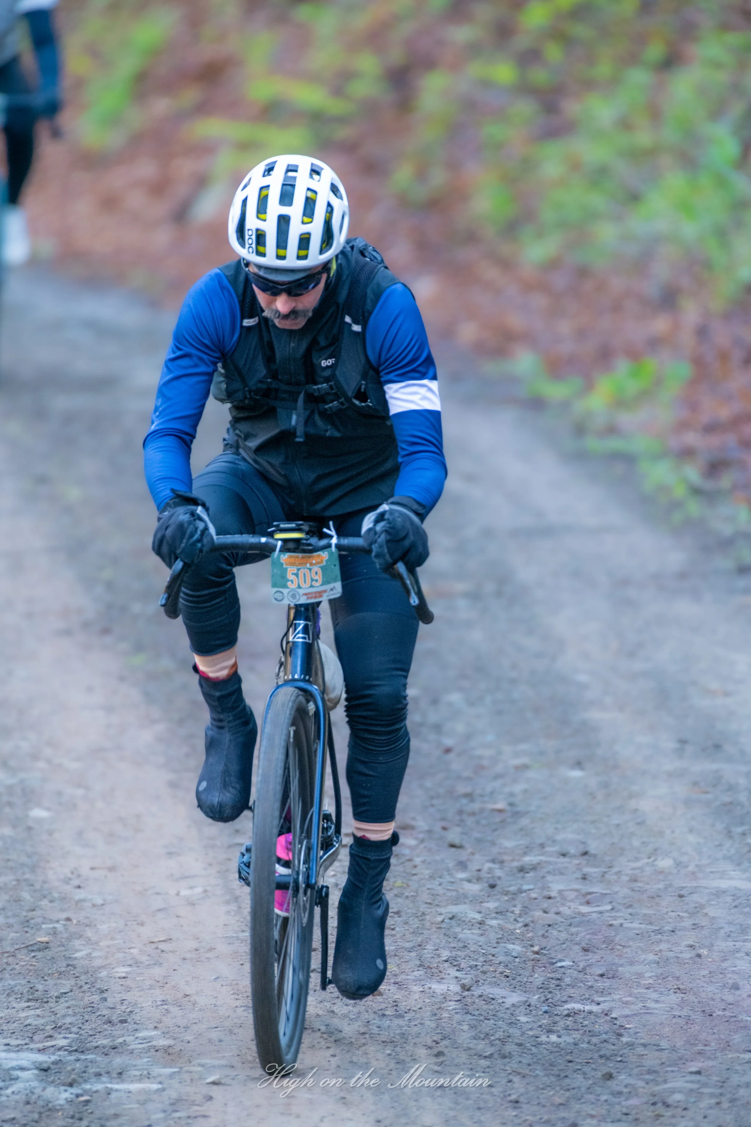 A person mountain biking on a dirt trail in a wooded area, wearing a helmet, sunglasses, and athletic gear.