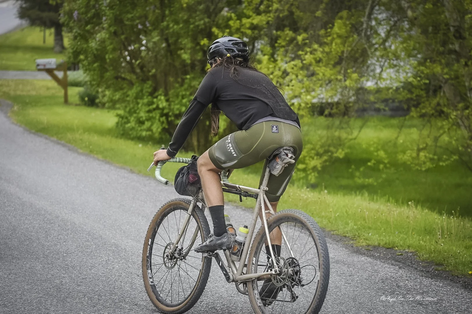 A person riding a gravel bike on a paved country road, wearing a helmet, black jersey, and green shorts, with a small saddlebag and water bottles, surrounded by greenery and trees.