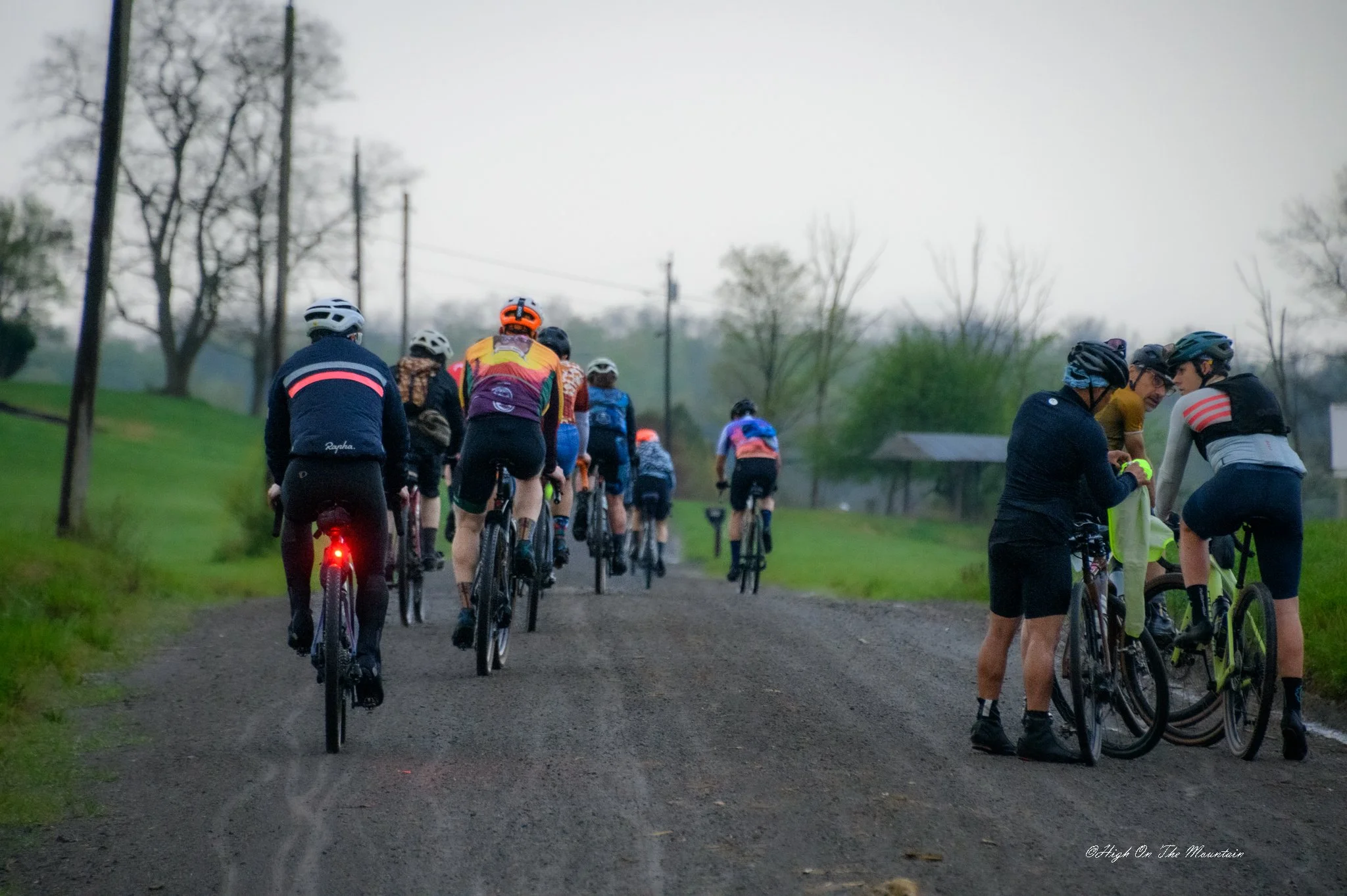A group of cyclists riding on a gravel road in a rural area during dusk or dawn, with some cyclists stopped on the side, adjusting their bikes or chatting.