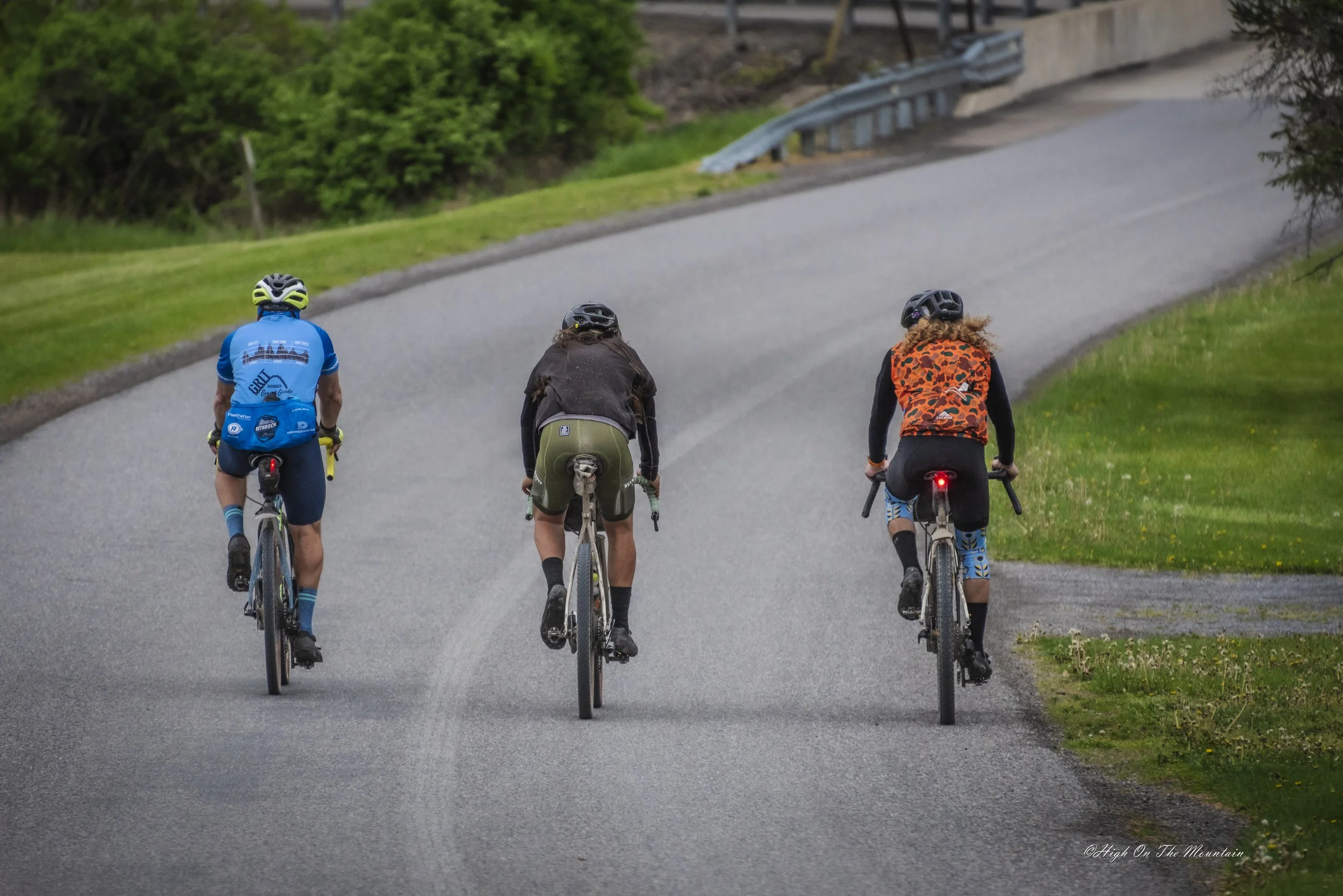 Three cyclists riding on a paved road with green grass and trees on either side, and a metal guardrail in the background.