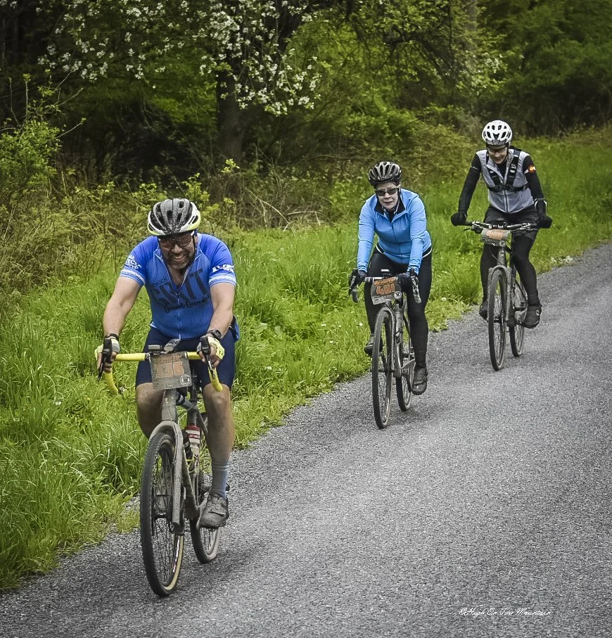 Three people riding bicycles on a gravel path surrounded by green trees and grass.