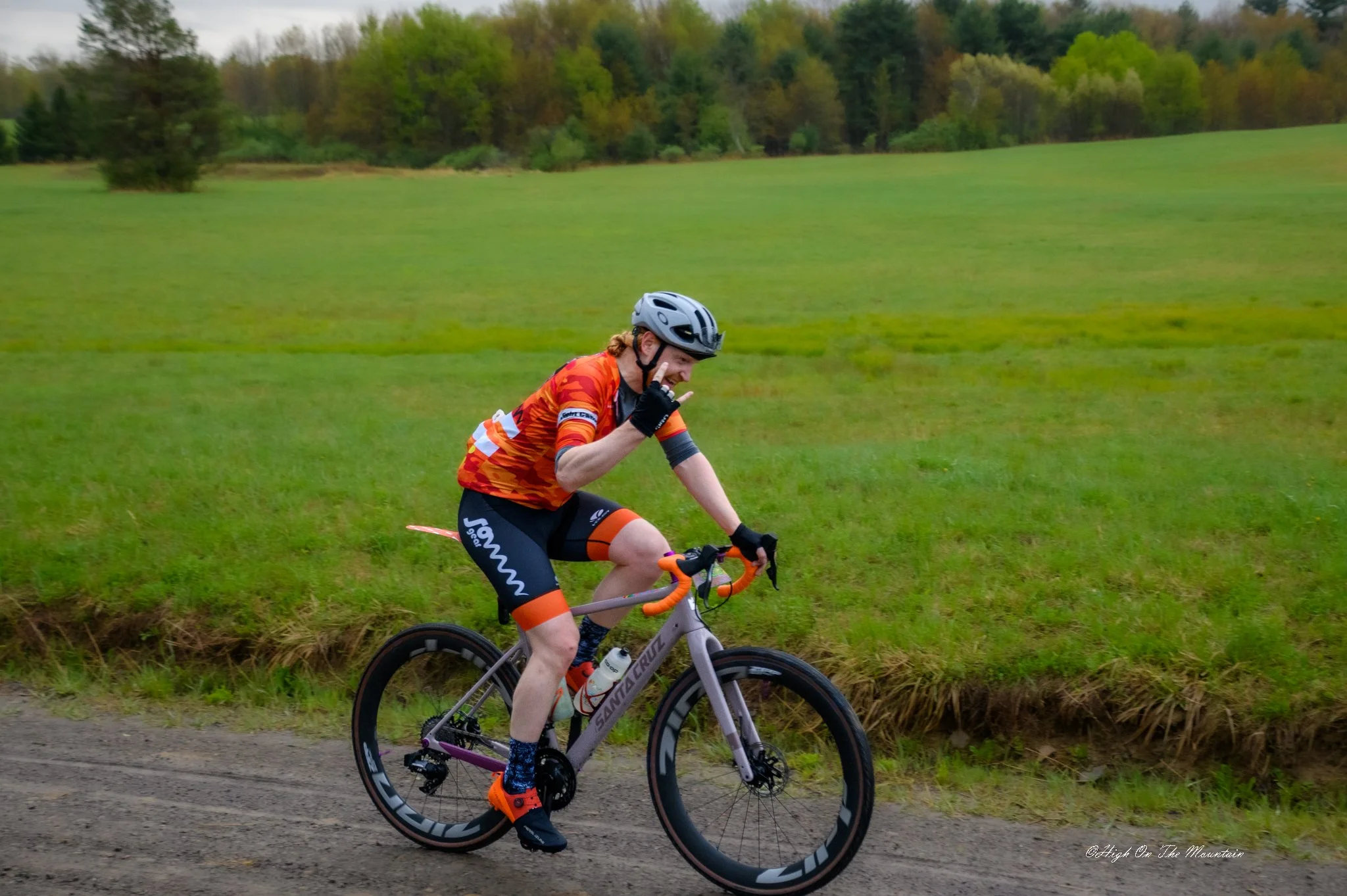 A person riding a bike on a dirt path near a grassy field with trees in the background, making a peace sign with their right hand and smiling, wearing a helmet and athletic clothing.