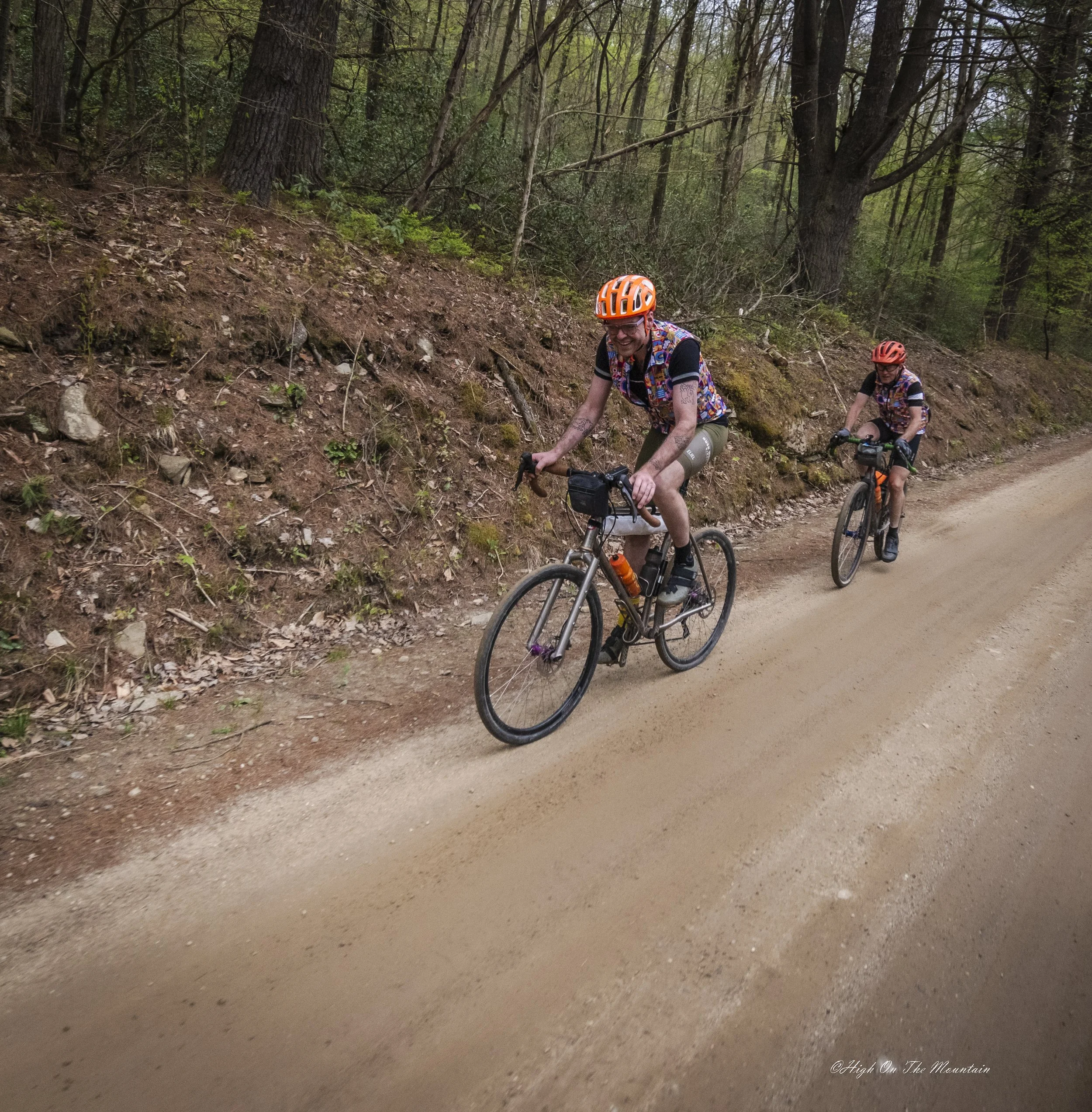 Two people mountain biking on a dirt trail through a forest with tall trees and green leaves, one wearing an orange helmet and colorful vest, smiling.