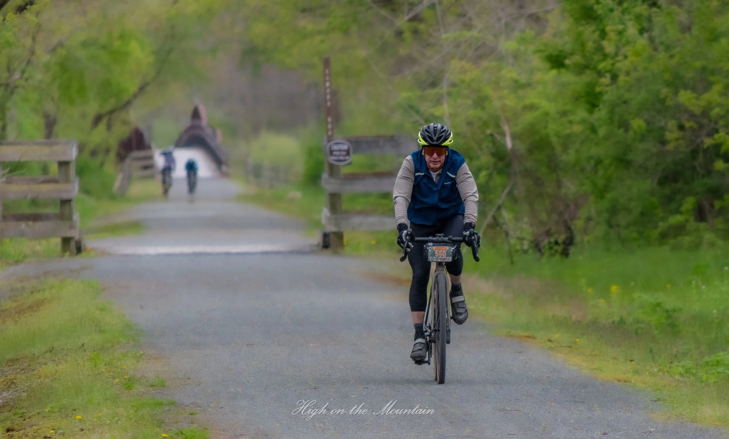 A man riding a bicycle on a paved trail surrounded by green trees and foliage. He wears a black helmet, sunglasses, and a blue vest. Two other people are visible in the background, walking along the trail. The scene is in a natural setting with a bri