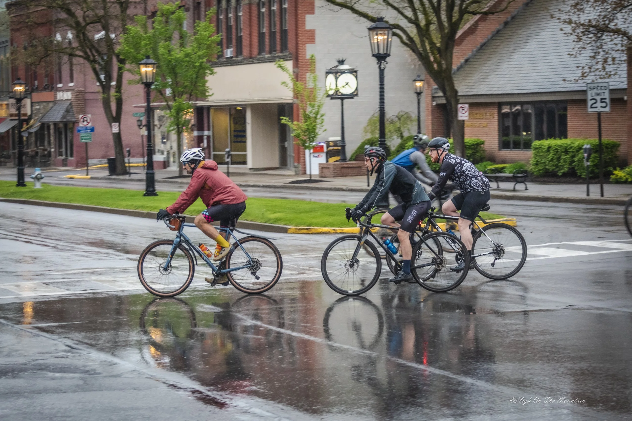 A group of cyclists riding their bikes on a wet street with puddles, in a city setting with brick and concrete buildings, trees, street lamps, and clock in the background during overcast weather.