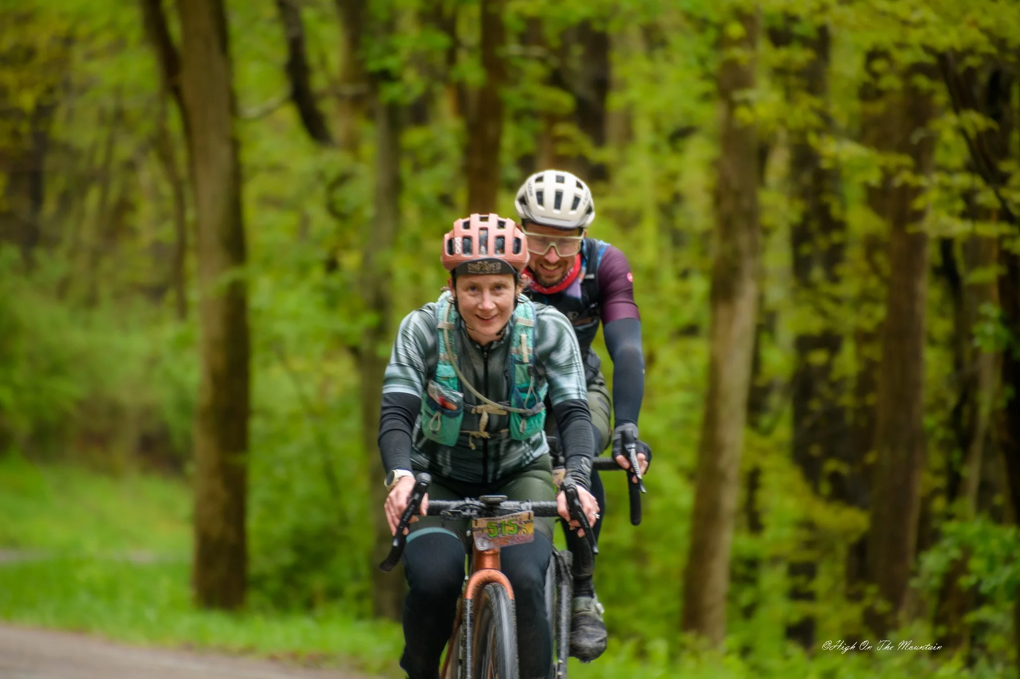 Two people riding mountain bikes through a forest with green trees, wearing helmets and athletic clothing, smiling and enjoying a sunny day.