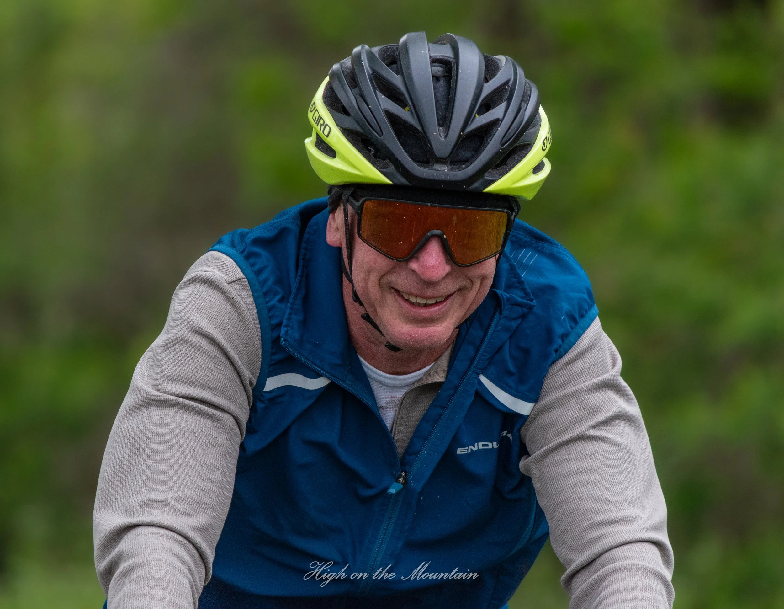 Close-up of a man wearing a black and yellow bike helmet, sunglasses, and a blue and gray jacket, riding a mountain bike outdoors surrounded by green trees.