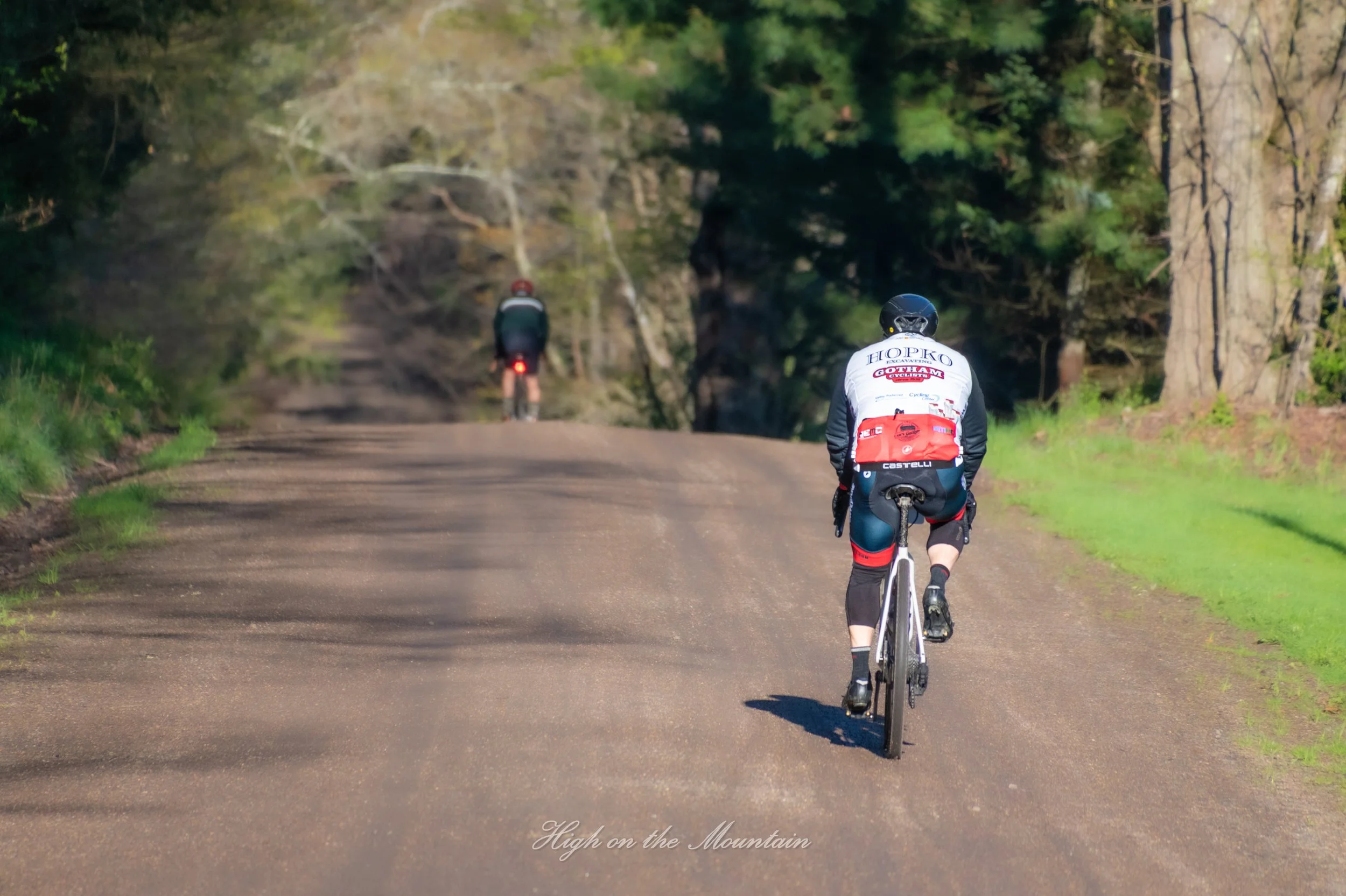 A person riding a bicycle on a dirt path through a wooded area, with another cyclist ahead in the distance. Trees line the sides of the trail. The photo has a text overlay at the bottom that says 'High on the Mountain.'