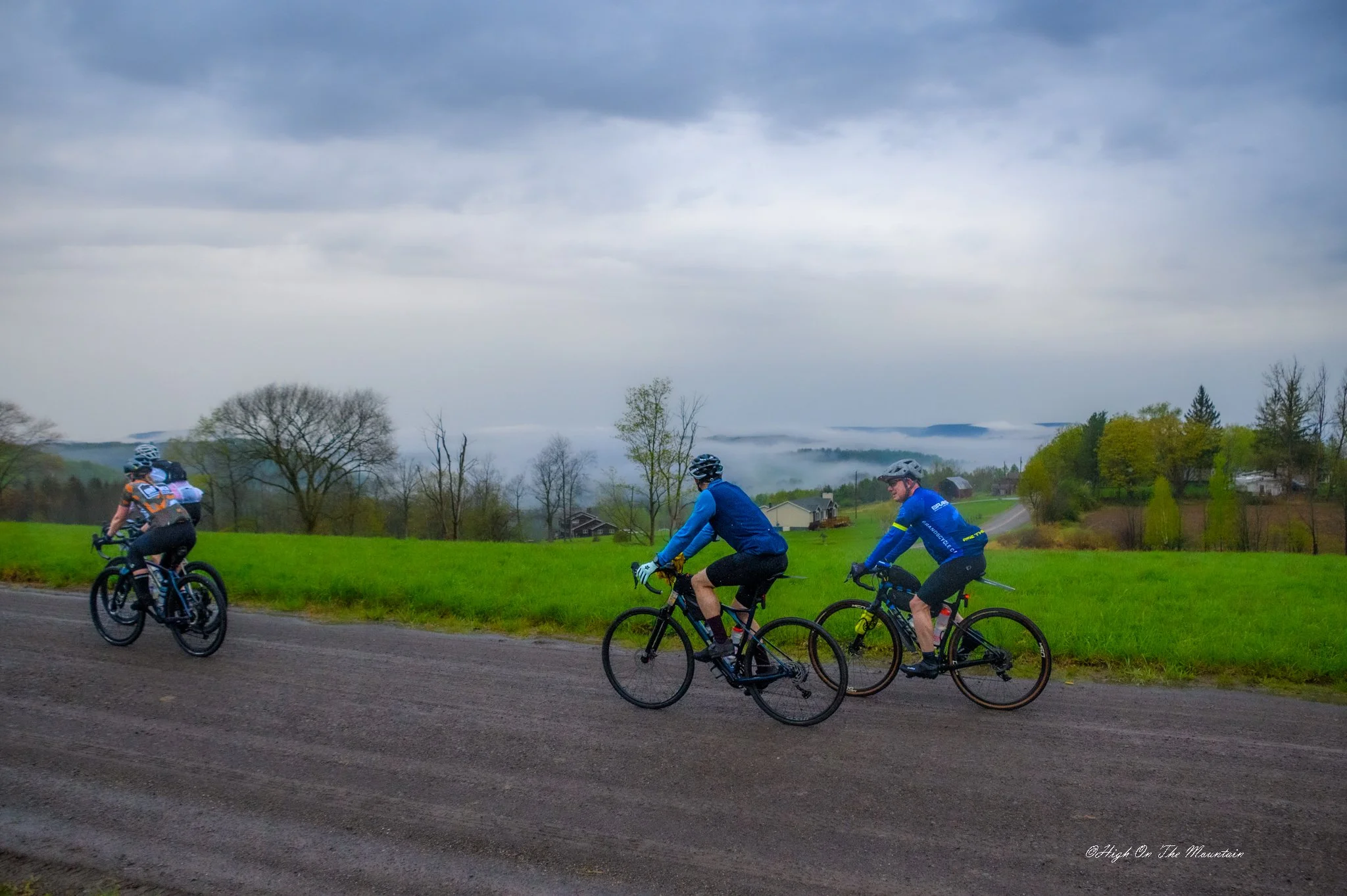 Group of cyclists riding on a paved path through an open area with trees, hills, and houses in the background on a cloudy day.