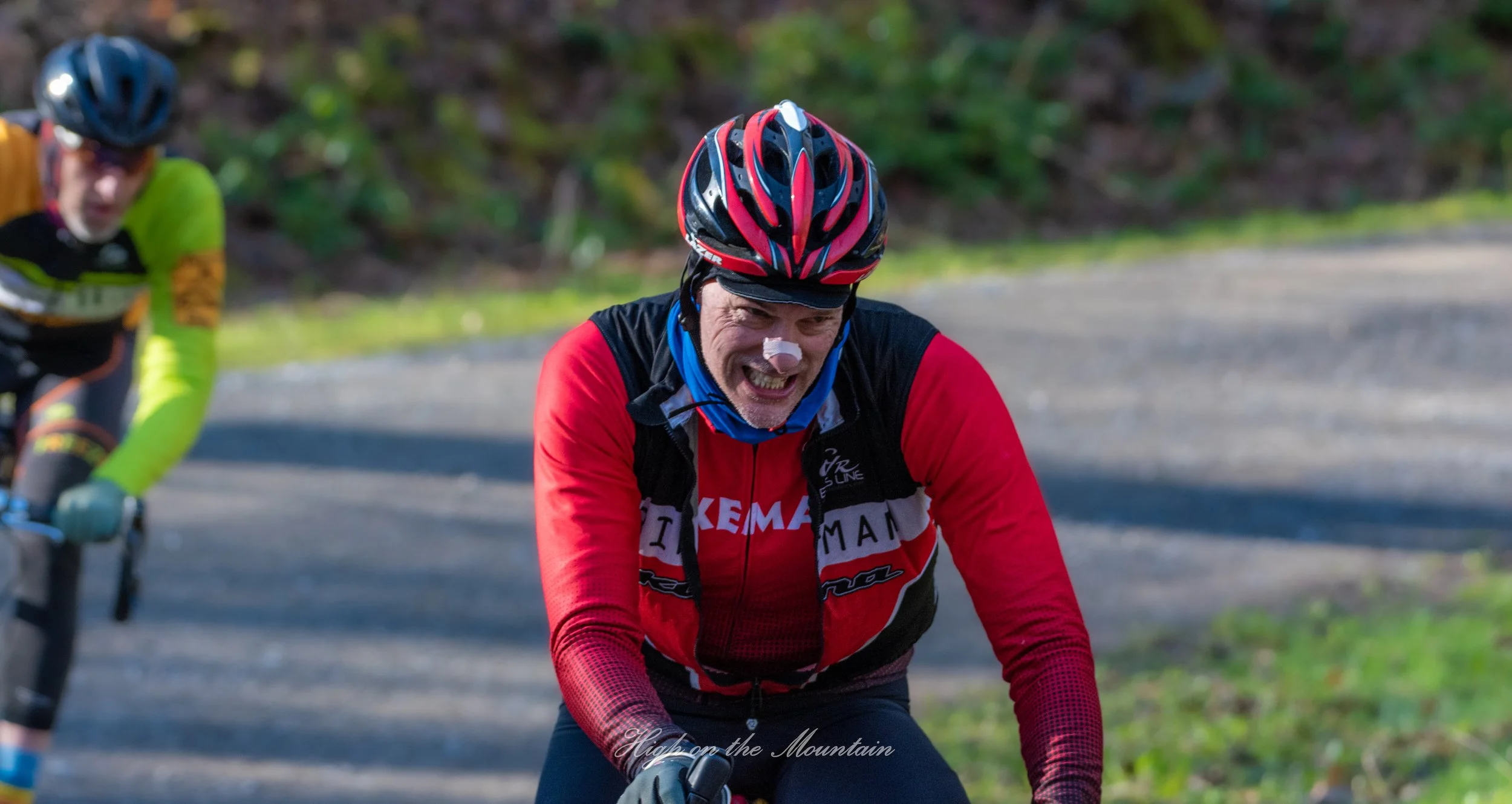 Two mountain bikers riding on a trail surrounded by greenery during daytime. The rider in the foreground is wearing a red and black helmet and a red jacket with black details, smiling and looking ahead. The rider in the background is wearing a blue h