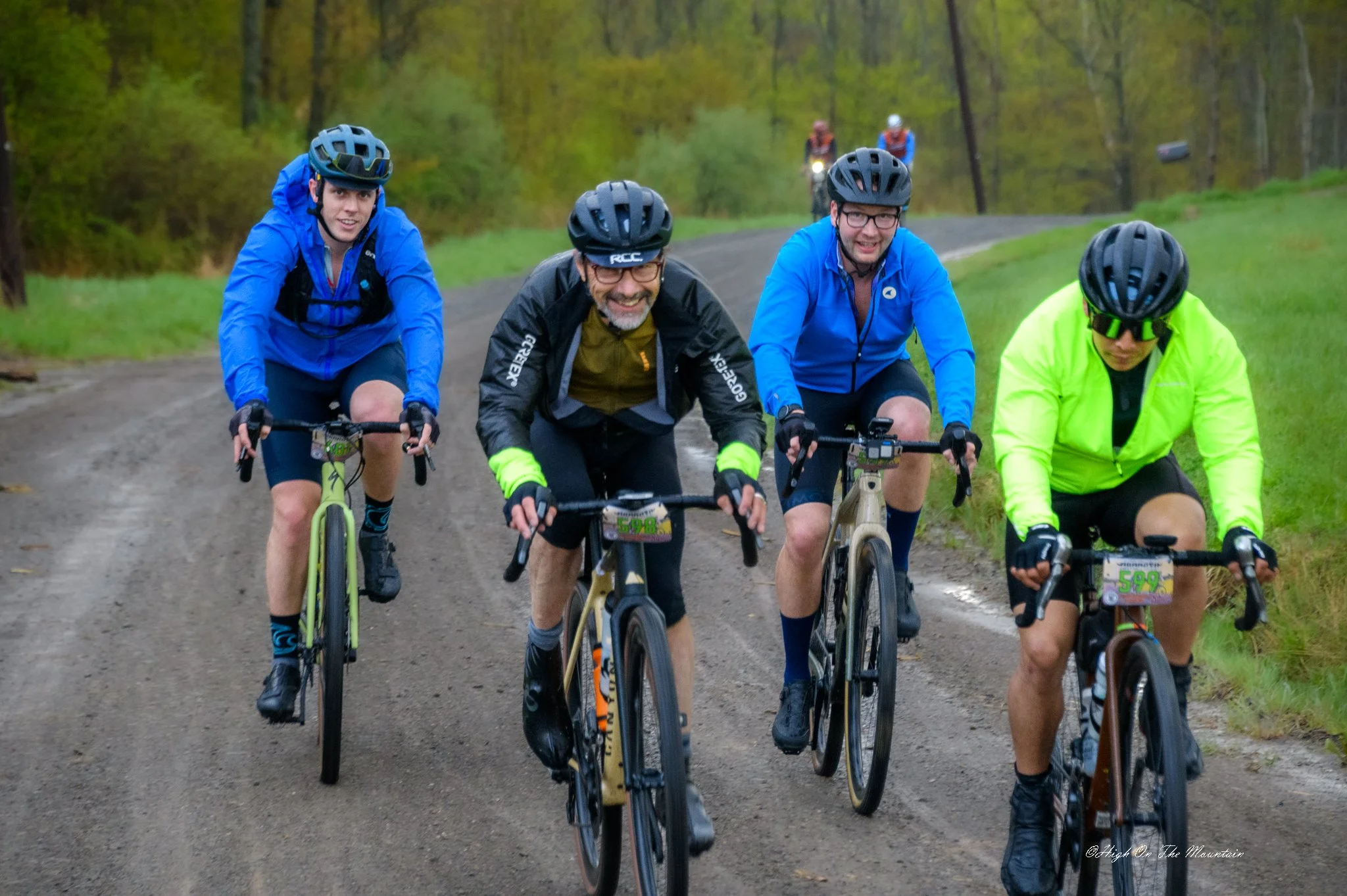 Group of five men riding bikes on a dirt trail in a wooded area with autumn foliage. They are wearing helmets and outdoor gear, smiling and focused on riding.