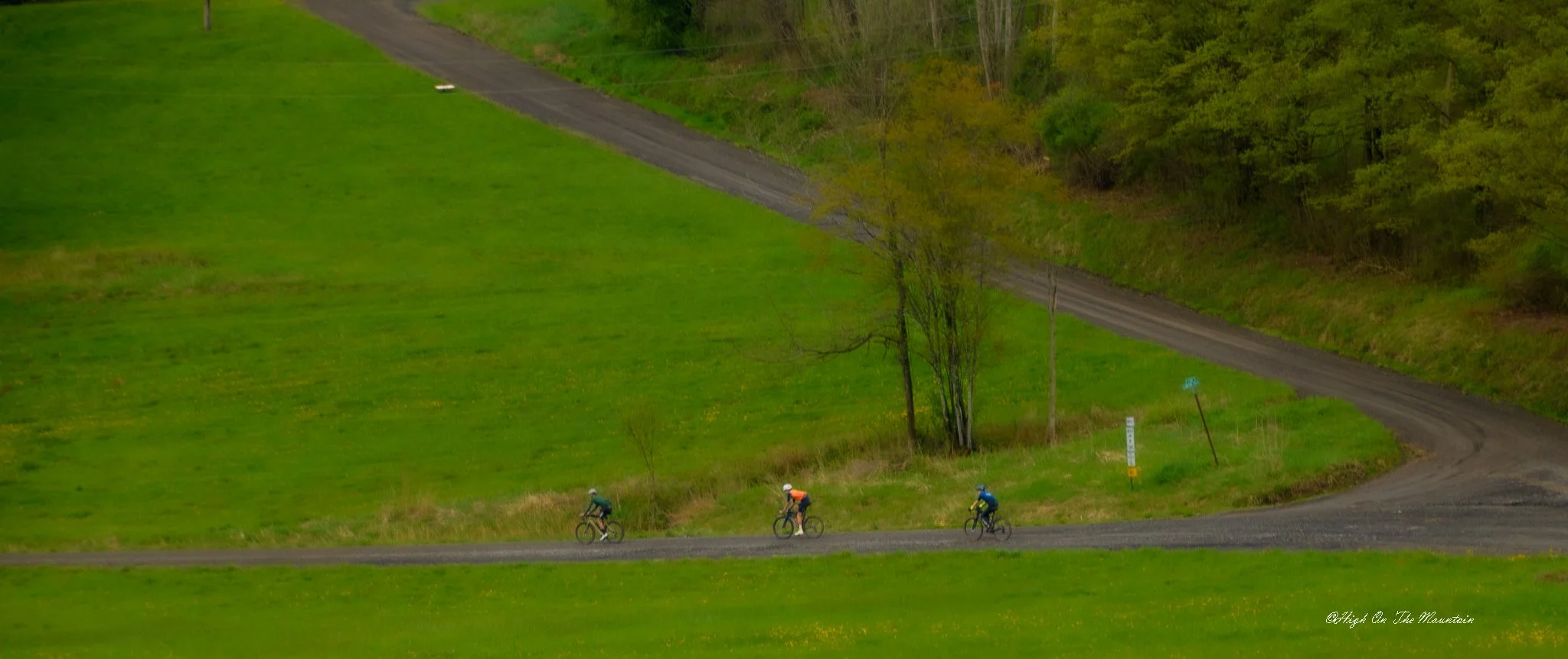 Three cyclists riding on a winding dirt trail through a lush green landscape with trees, grassy fields, and hills.