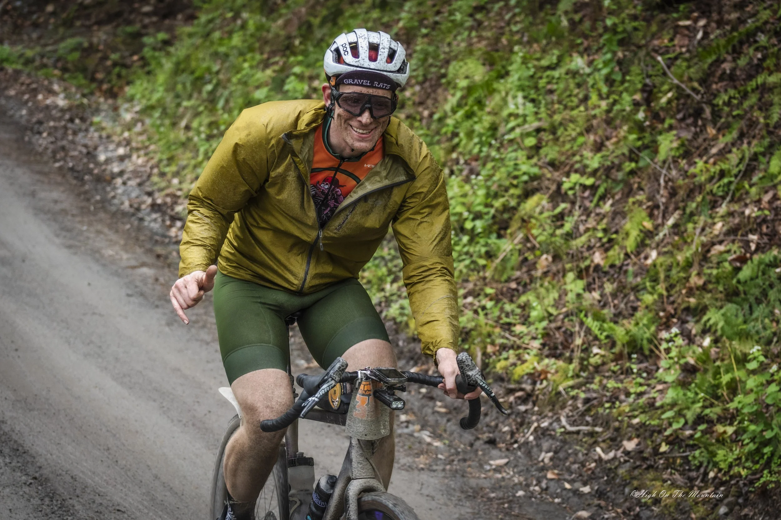 A man riding a mountain bike on a dirt trail through a forest, smiling, wearing a white helmet, sunglasses, a yellow jacket, orange shirt, and green shorts.