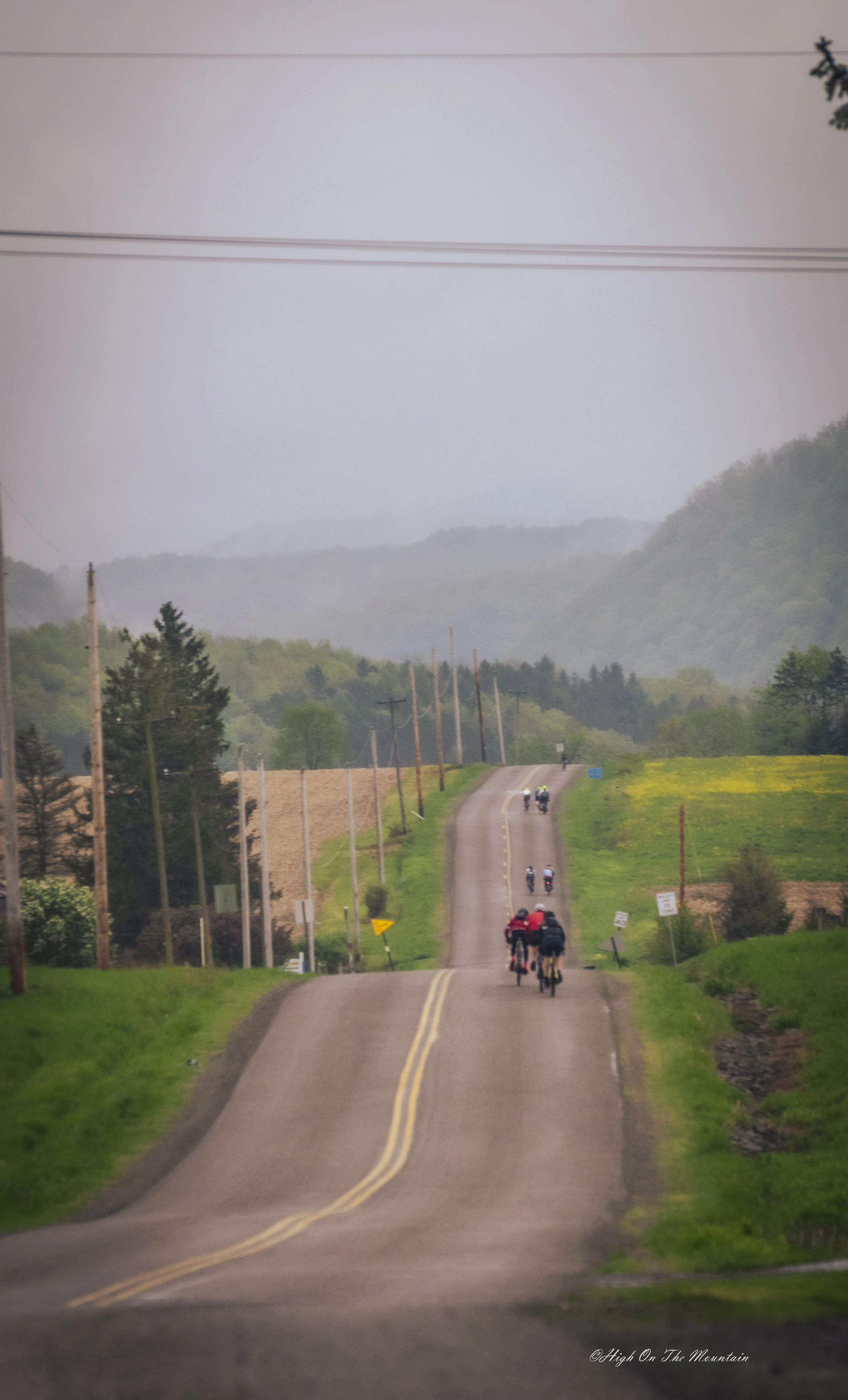 Long, winding rural road with yellow dividing lines, flanked by green grass and trees, hills in the background, and several people riding bikes along the road on a misty day.