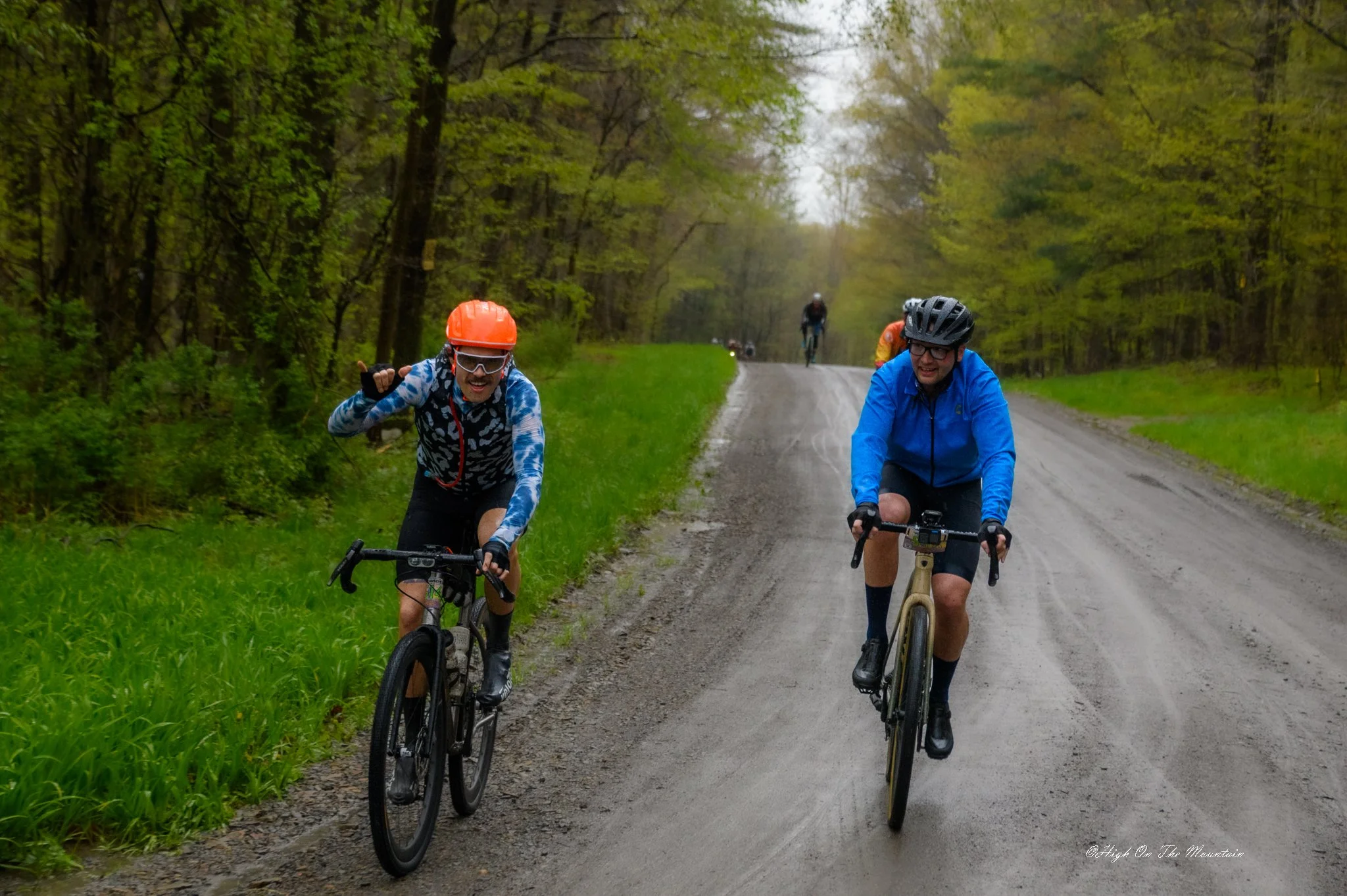 Two men riding mountain bikes on a dirt trail through a green forest, wearing helmets and outdoor clothing, with more cyclists in the background.