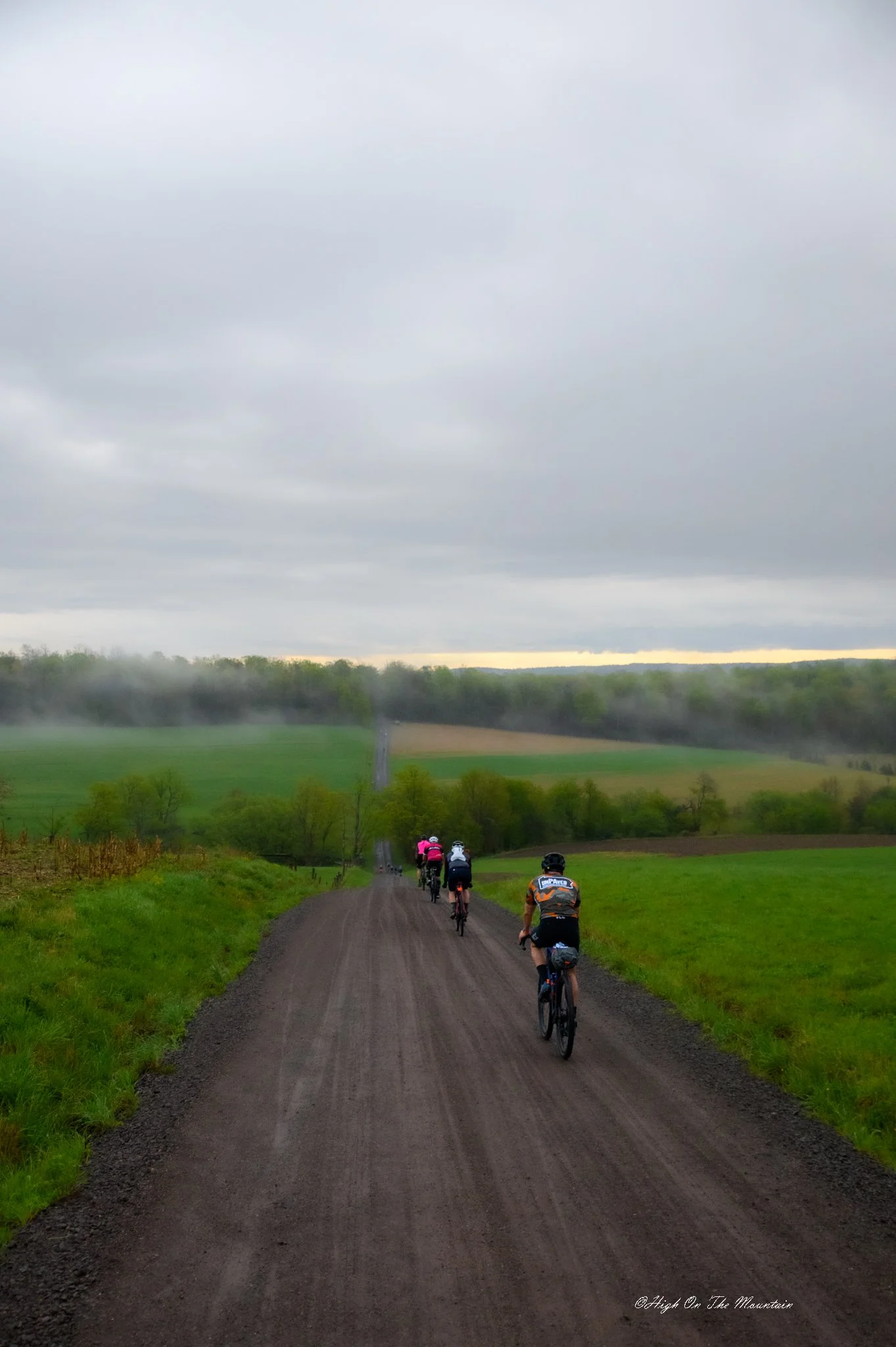 A group of cyclists riding on a dirt trail through a scenic, rural landscape with green fields, trees, and cloudy sky.