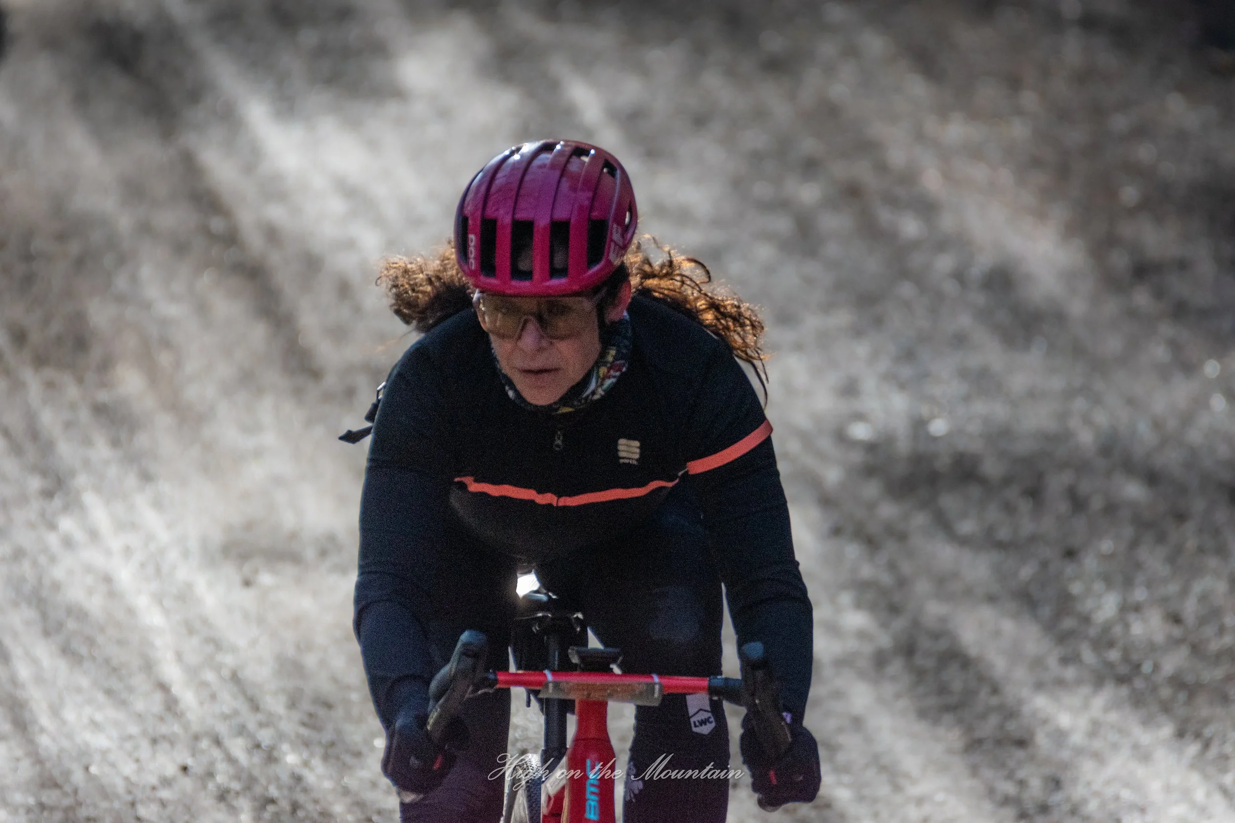 A woman riding a mountain bike on a rocky trail, wearing a pink helmet, glasses, and a black jacket with pink accents.