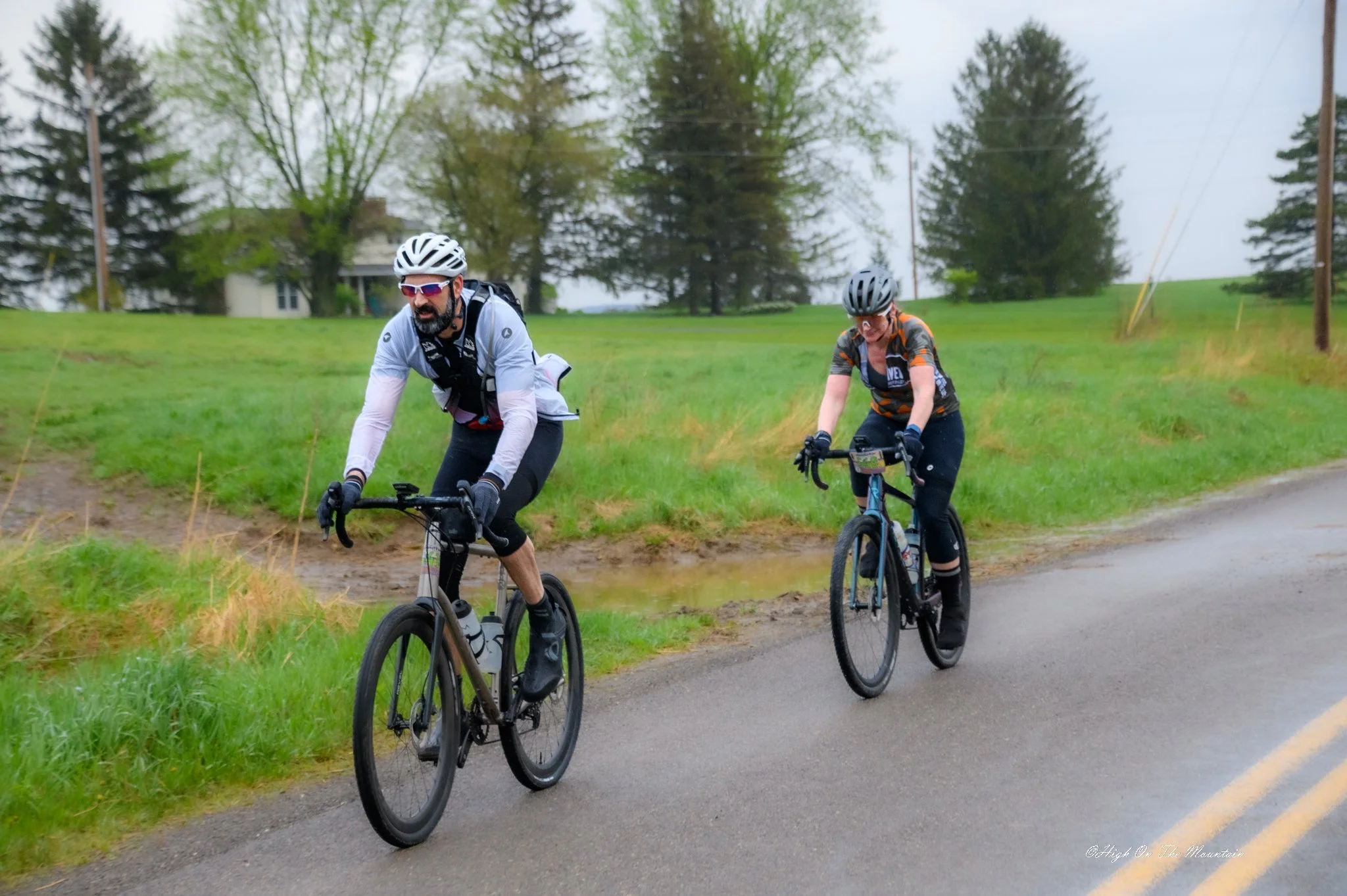 Two cyclists riding on a rural paved road with grassy fields, trees, and power lines in the background on a cloudy day.