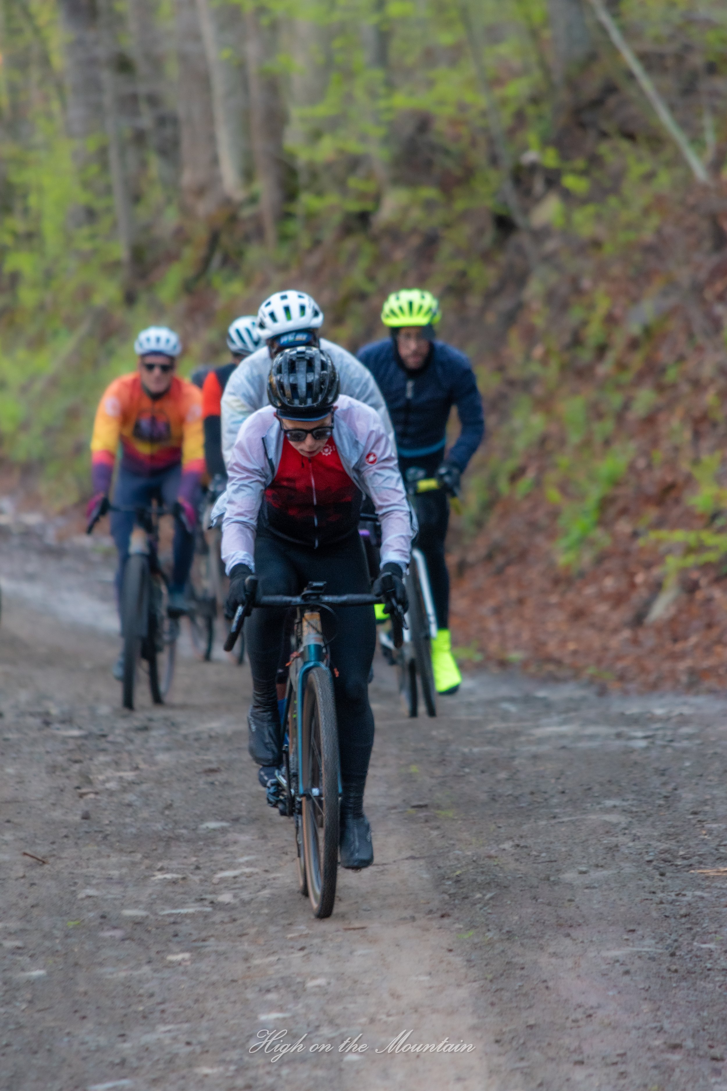 A group of cyclists riding mountain bikes on a dirt trail through a wooded area with green trees and foliage.