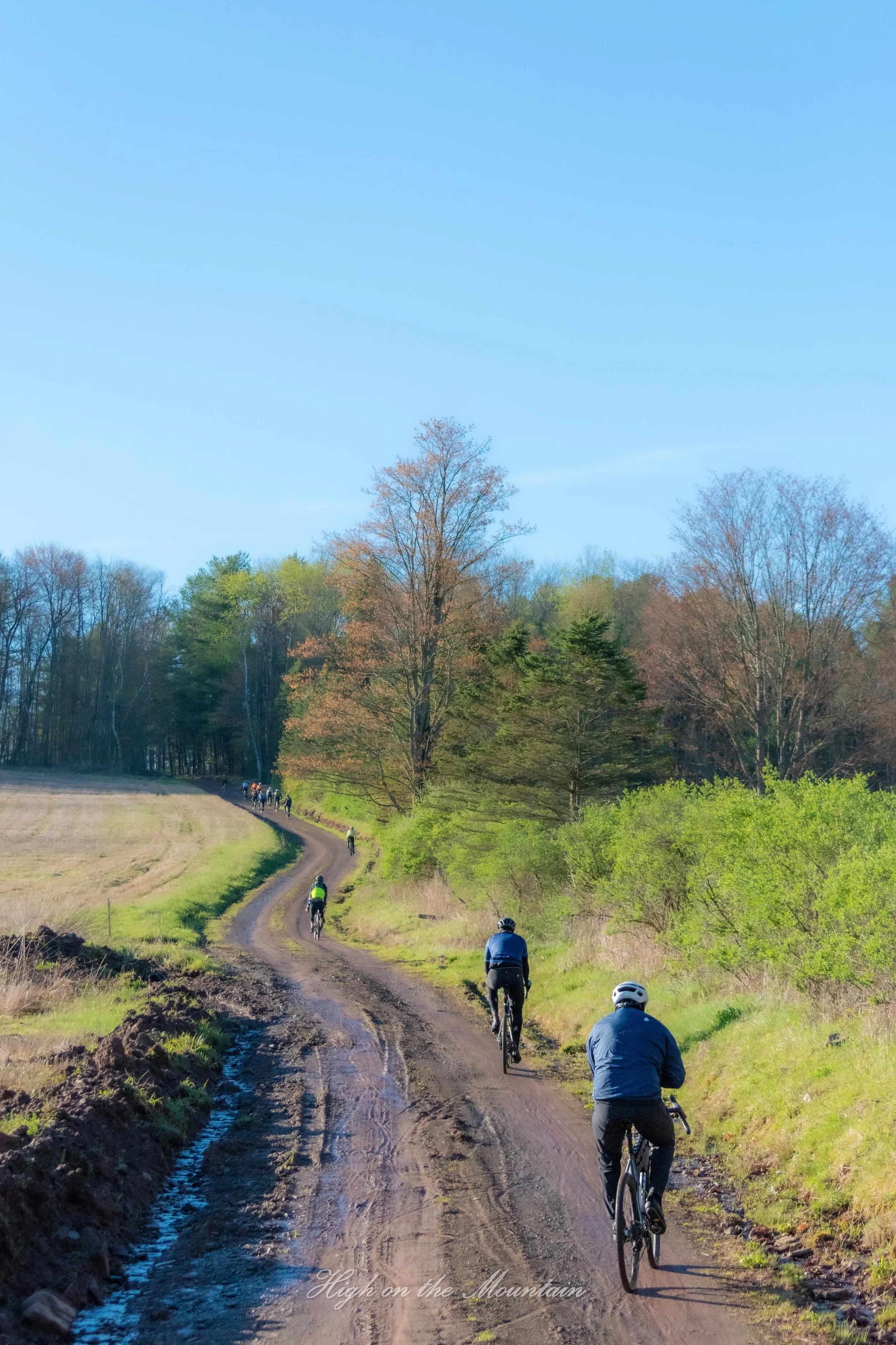 A group of cyclists riding on a dirt trail surrounded by trees with green and brown foliage under a blue sky. The trail is uneven with patches of mud and water, and the cyclists are wearing helmets and outdoor gear. The image has a text overlay at th