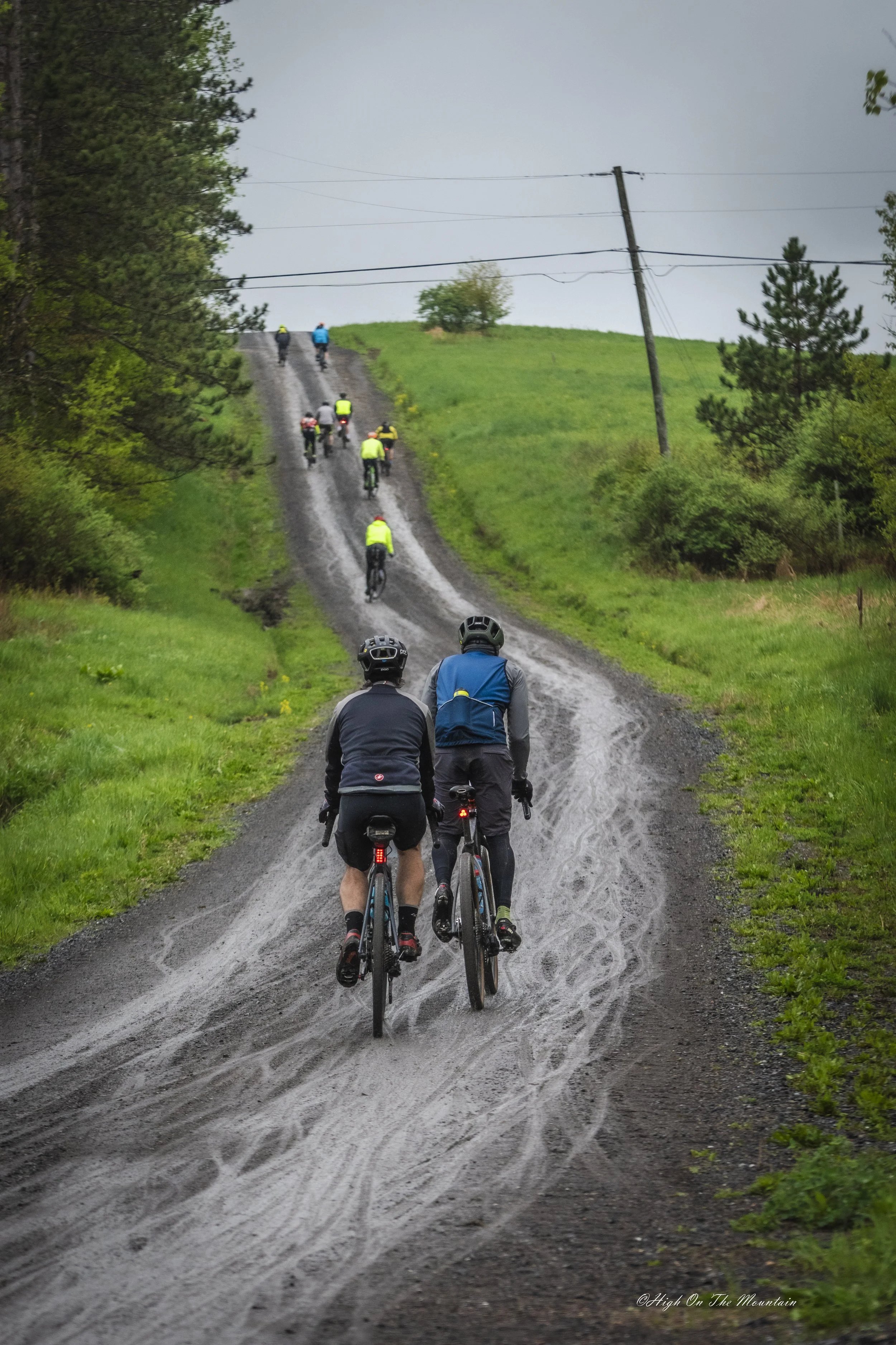 A group of cyclists riding up a muddy, dirt trail surrounded by green trees and grass, under cloudy skies.