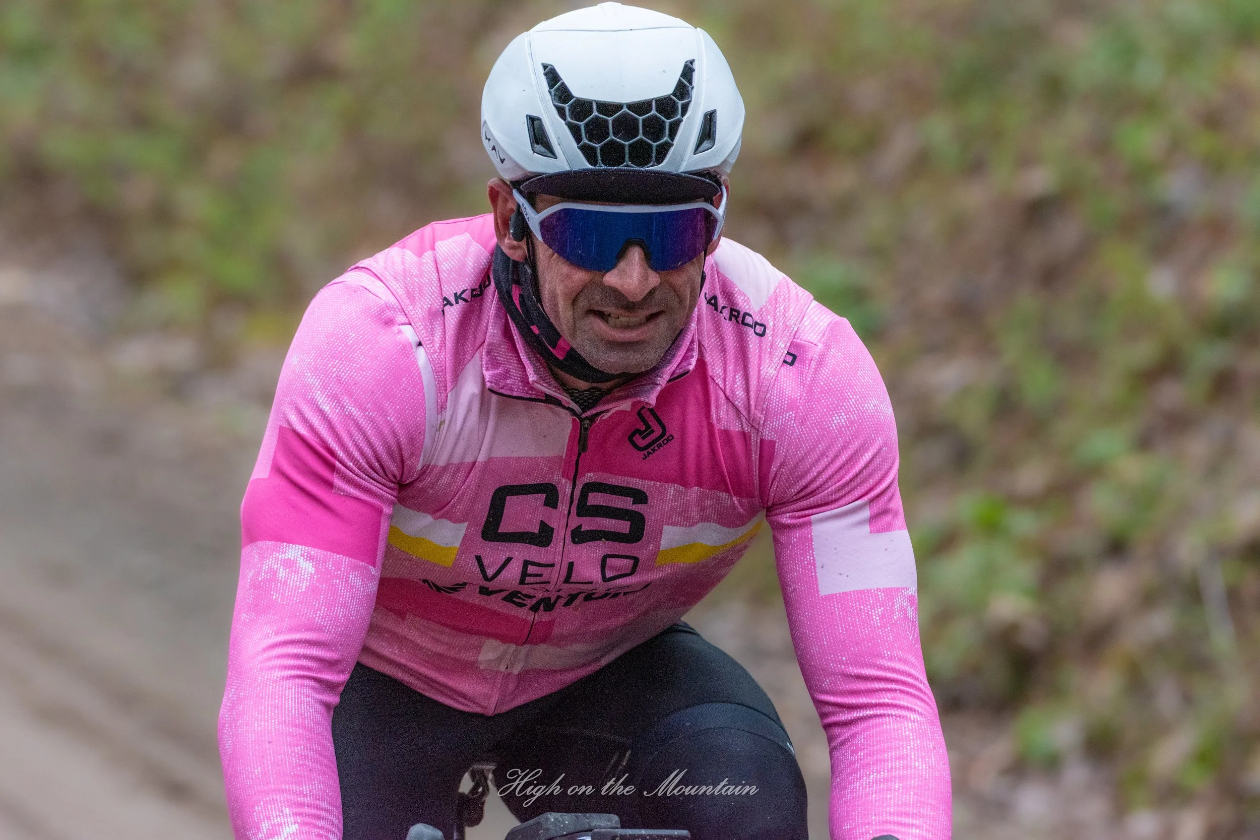 Cyclist wearing a pink jersey and a white helmet with black honeycomb pattern, riding on a trail with blurred greenery in the background.