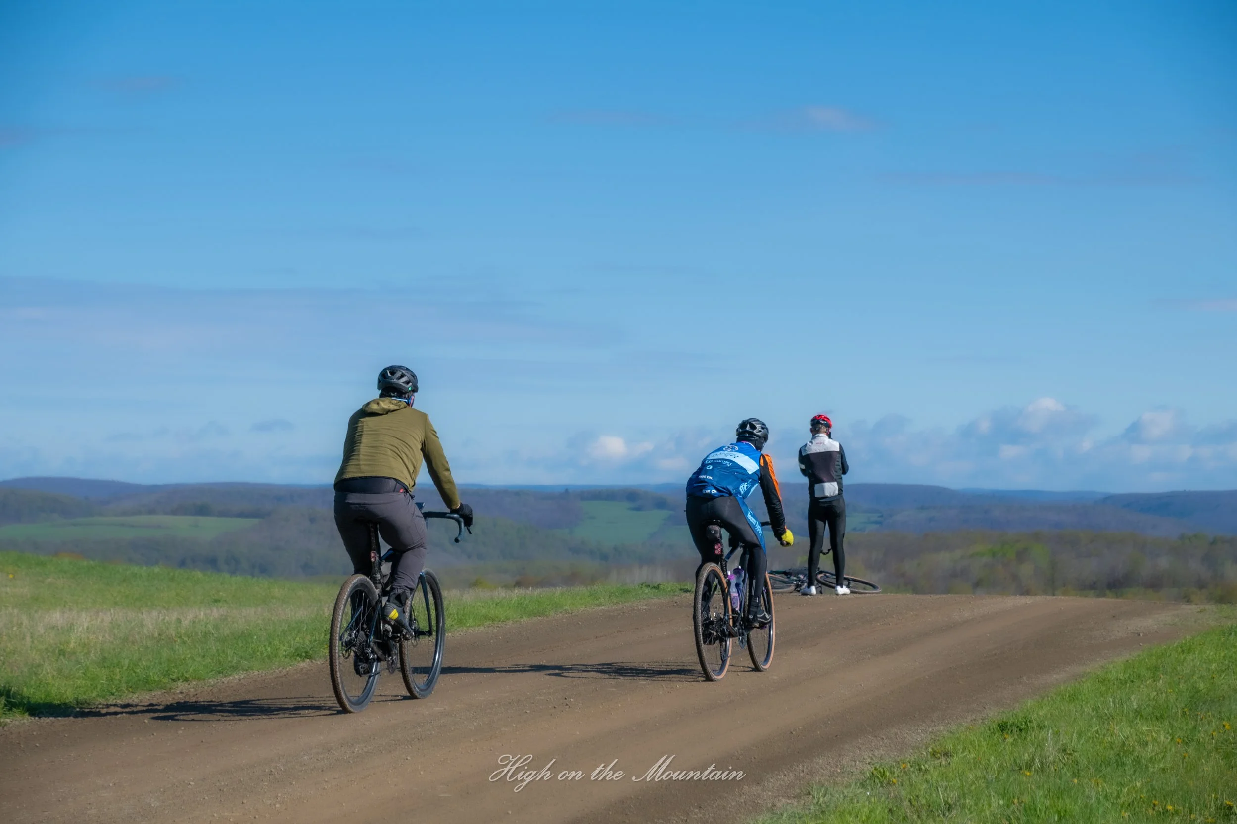 Four cyclists riding on a dirt path through a grassy landscape under a clear blue sky, with rolling hills in the distance.