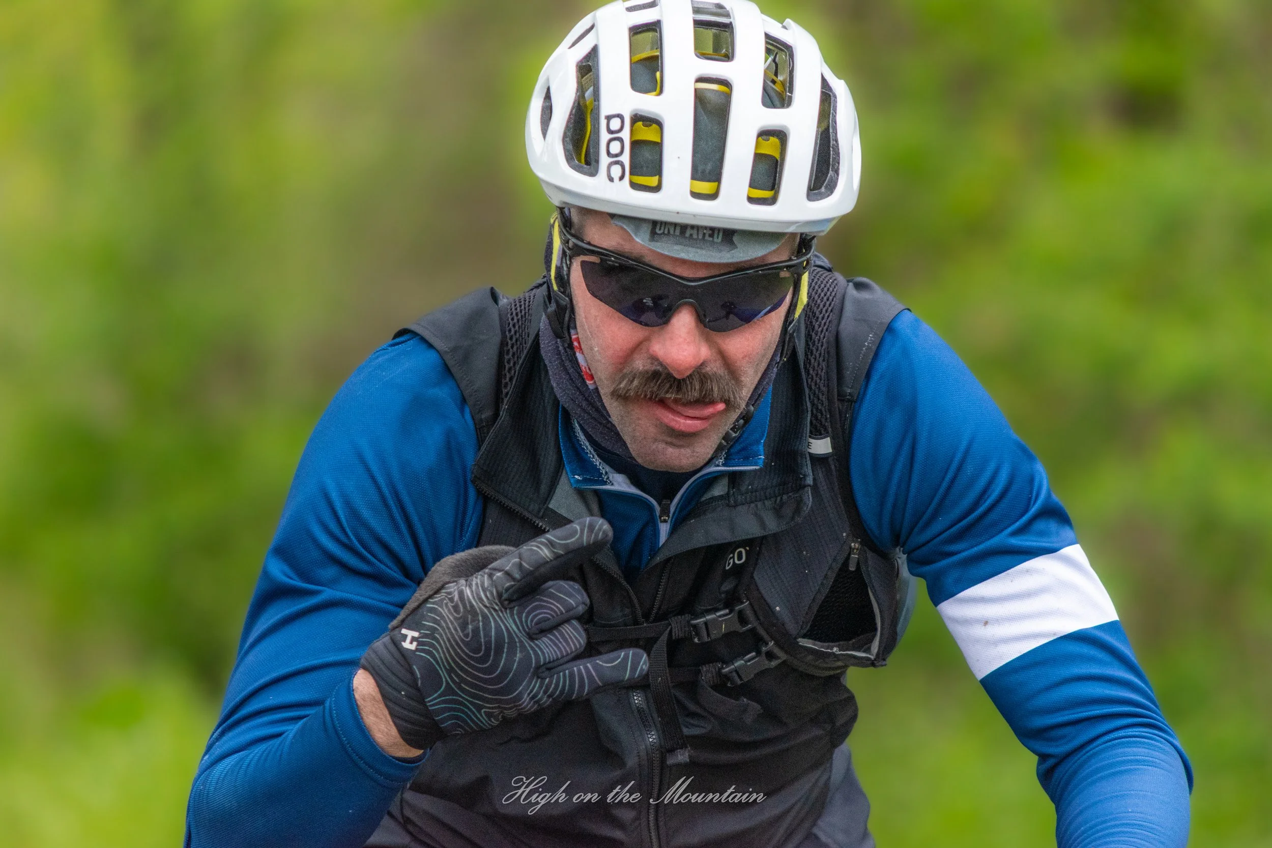 A man wearing a white helmet, black sunglasses, blue and black motorcycle gear, and gray gloves riding a mountain bike outdoors with a blurred green background.