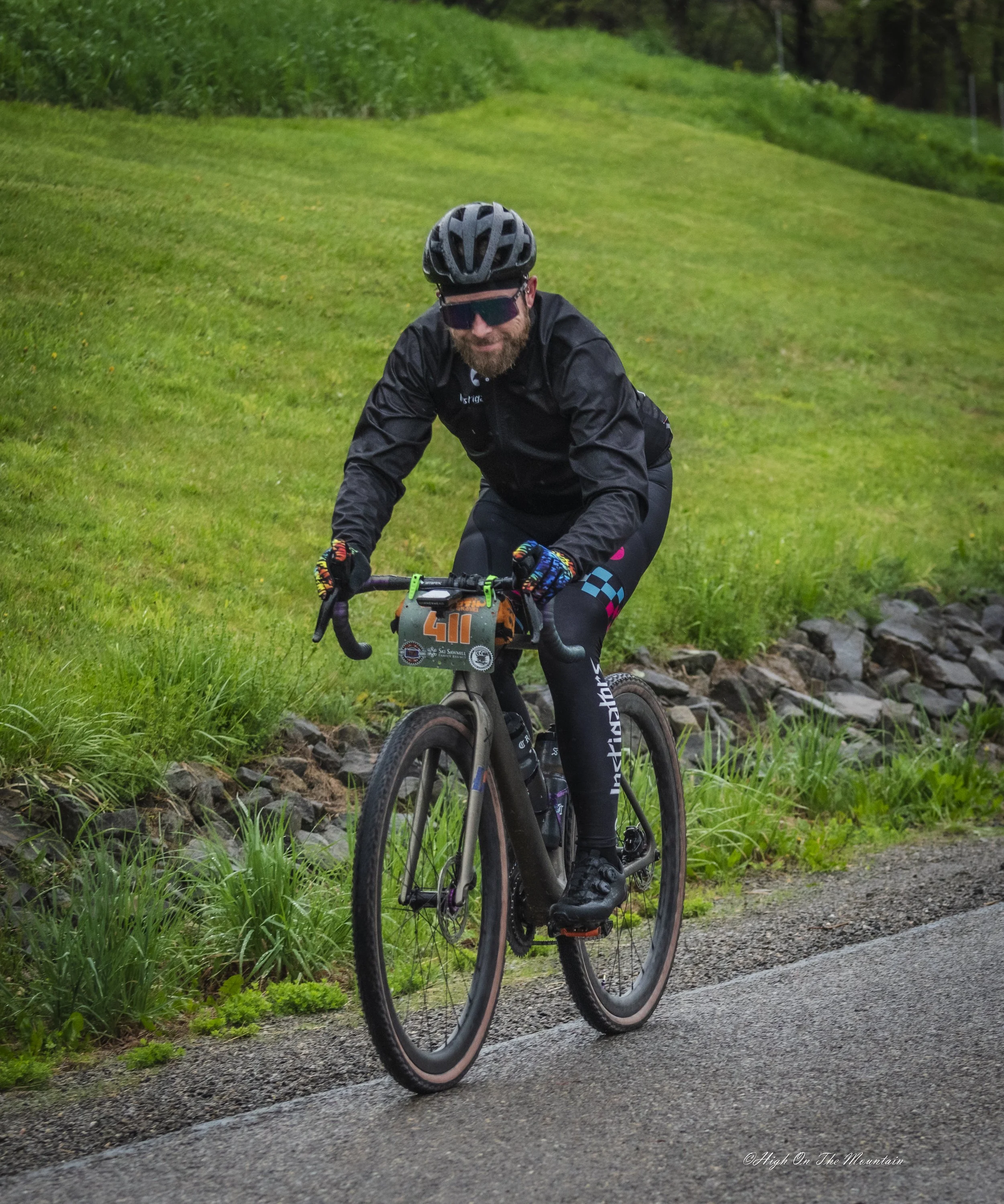 A cyclist with a helmet and sunglasses riding a mountain bike on a gravel path next to green grass and vegetation.