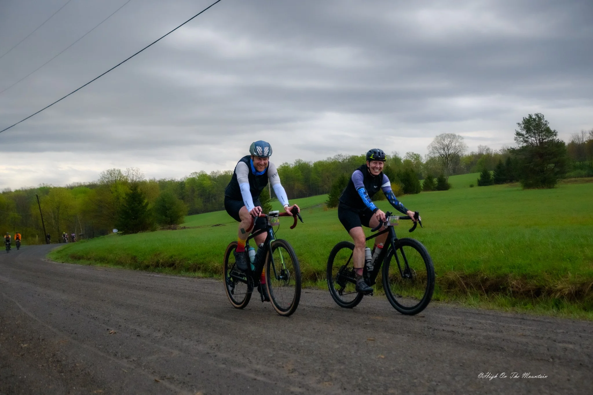 Two people riding bikes on a rural road with green fields and trees, cloudy sky overhead.