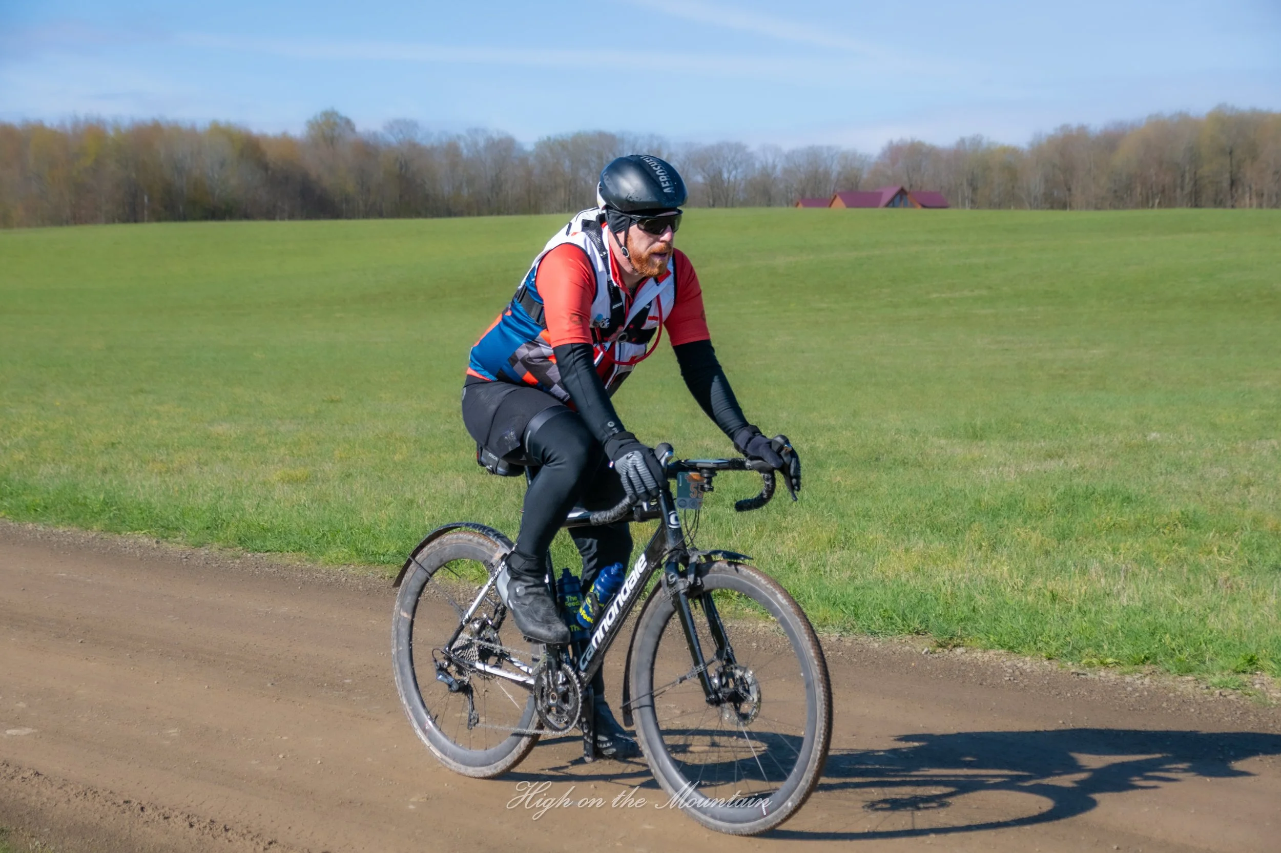 A man riding a mountain bike on a dirt trail in a rural area with green fields and trees in the background, wearing a helmet, sunglasses, and athletic clothing.