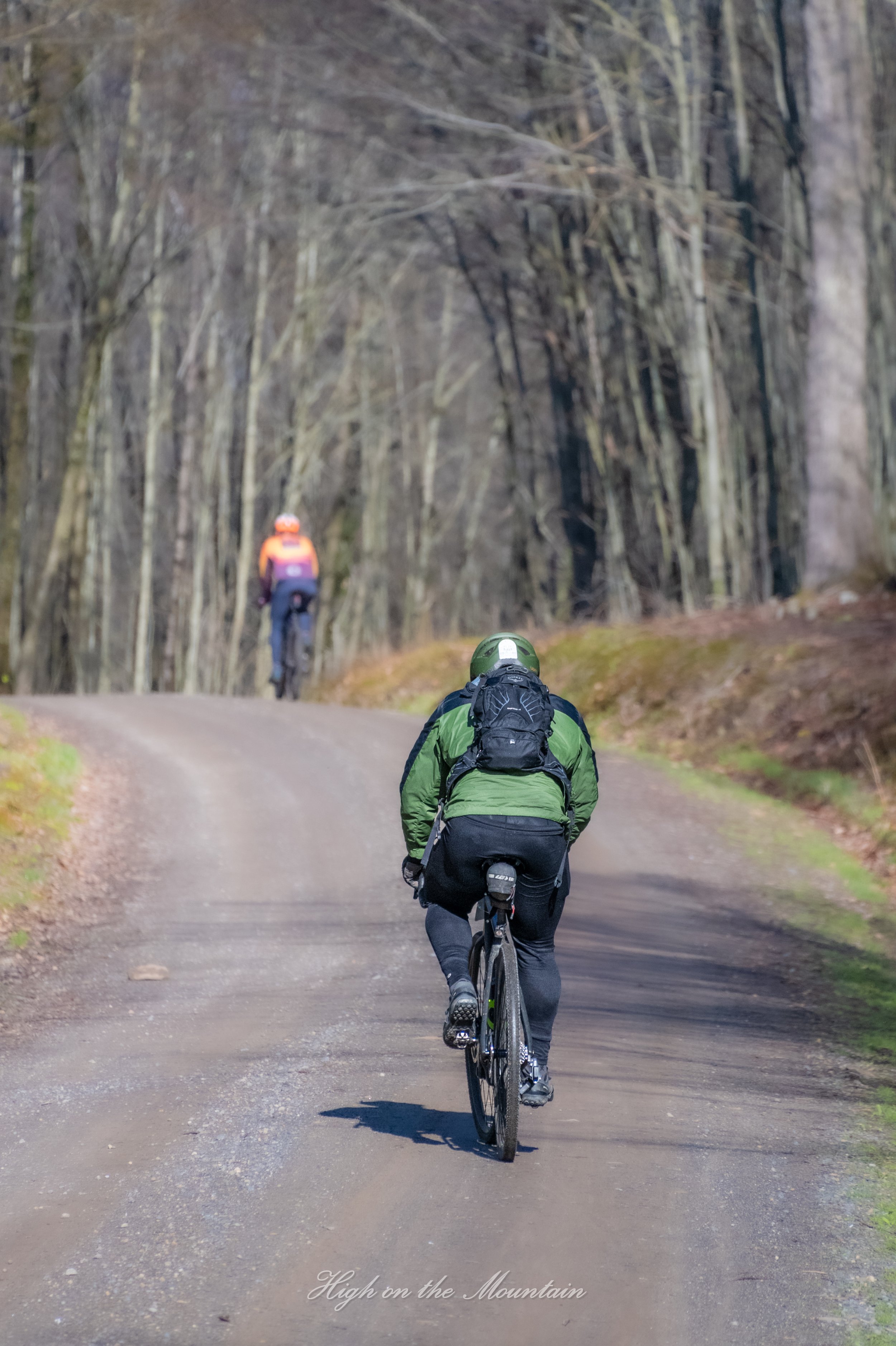 A person riding a bicycle on a dirt trail in a wooded area with leafless trees during the day. Another cyclist in an orange jacket is visible ahead on the trail. The text "High on the Mountain" is overlaid at the bottom of the image.
