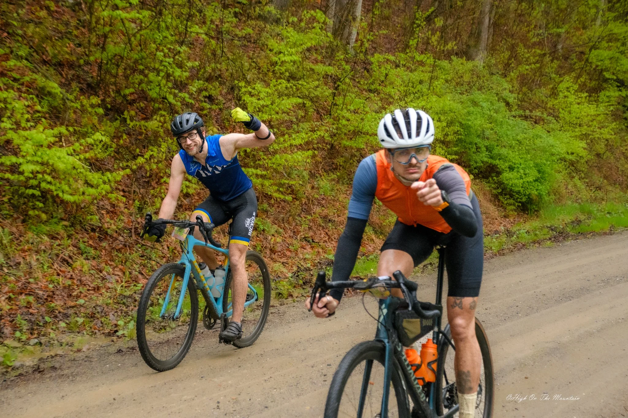 Two male mountain bikers on a dirt trail surrounded by green foliage. One is wearing a black helmet and glasses, smiling and waving. The other is wearing a white helmet and orange vest, focused on riding.
