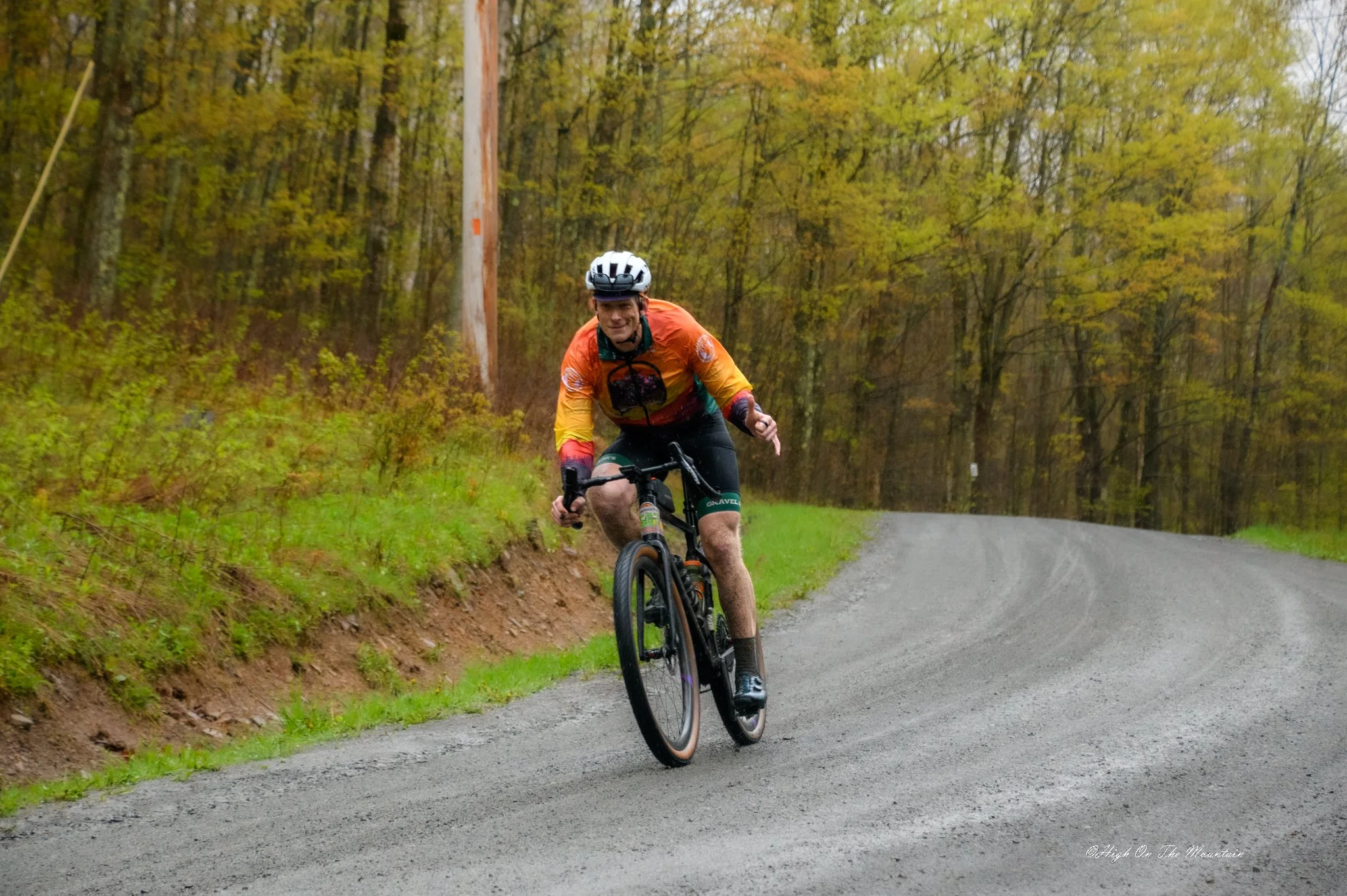 A man wearing a helmet and colorful bike jersey riding a mountain bike on a dirt trail through a forest with green trees in autumn.