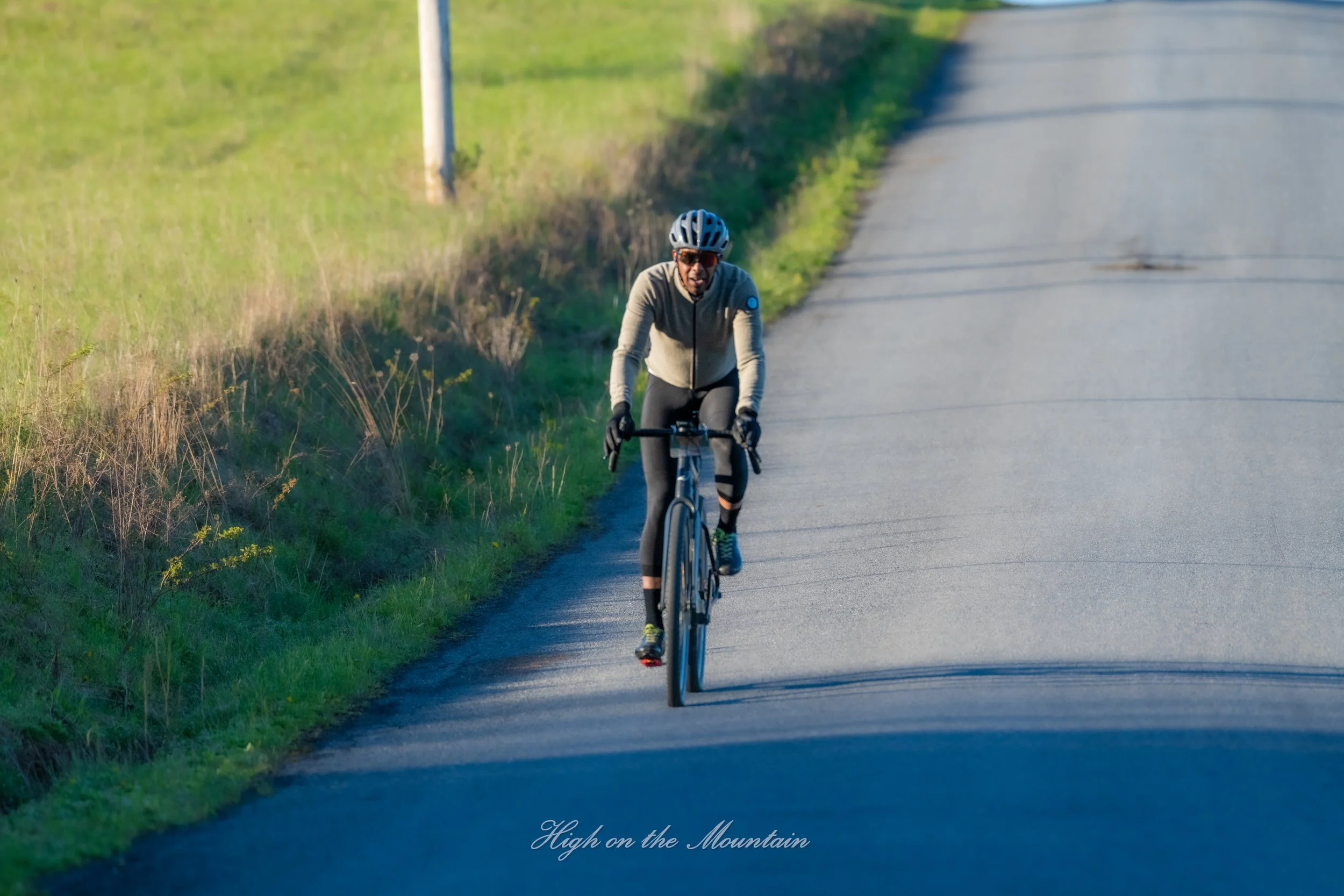 A cyclist riding on a country road during daylight. The cyclist wears a helmet, sunglasses, and athletic clothing. Green grass and a utility pole are visible along the side of the road. The scene has a peaceful, rural atmosphere.