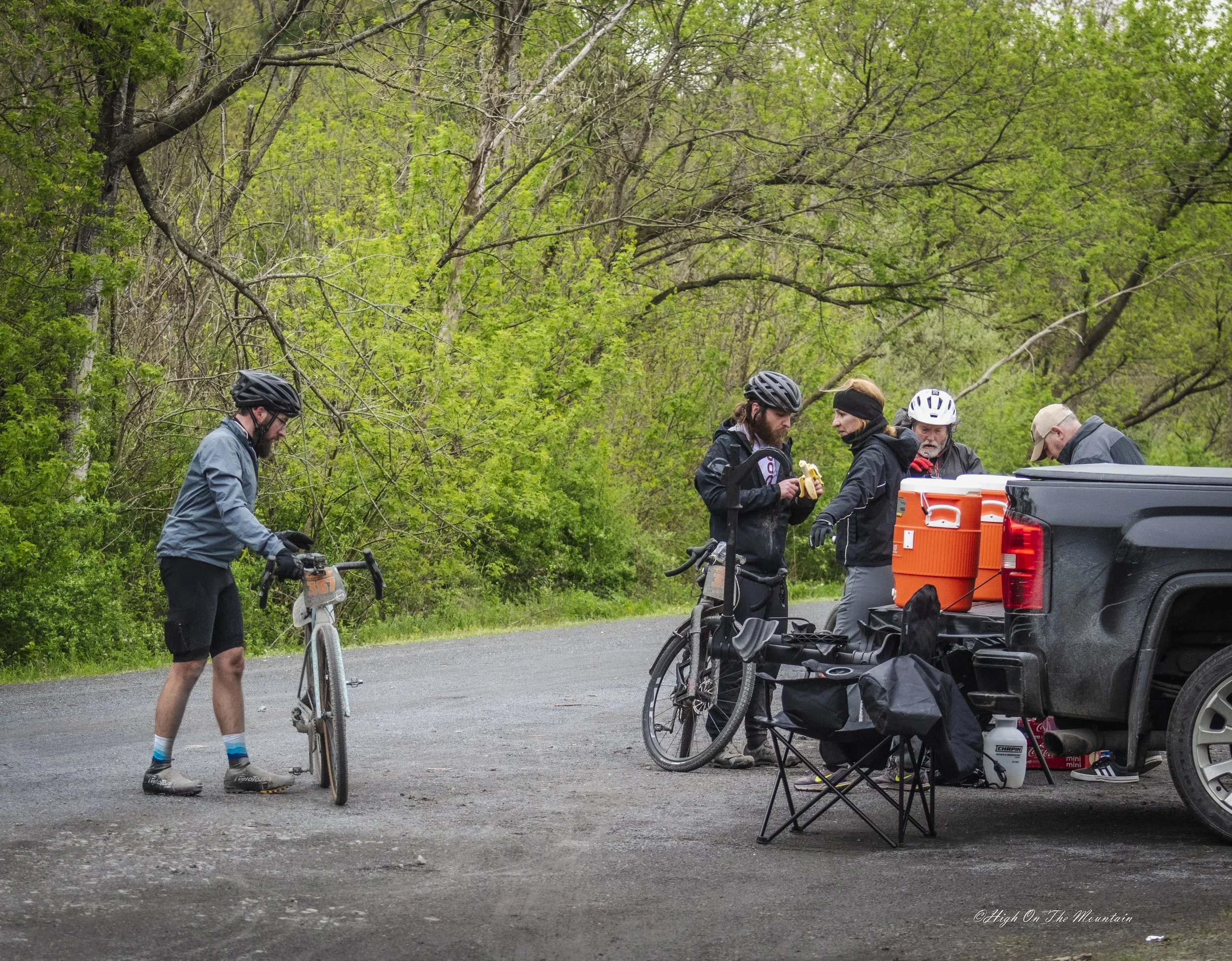 Group of five cyclists preparing at the back of a pickup truck in a wooded area.