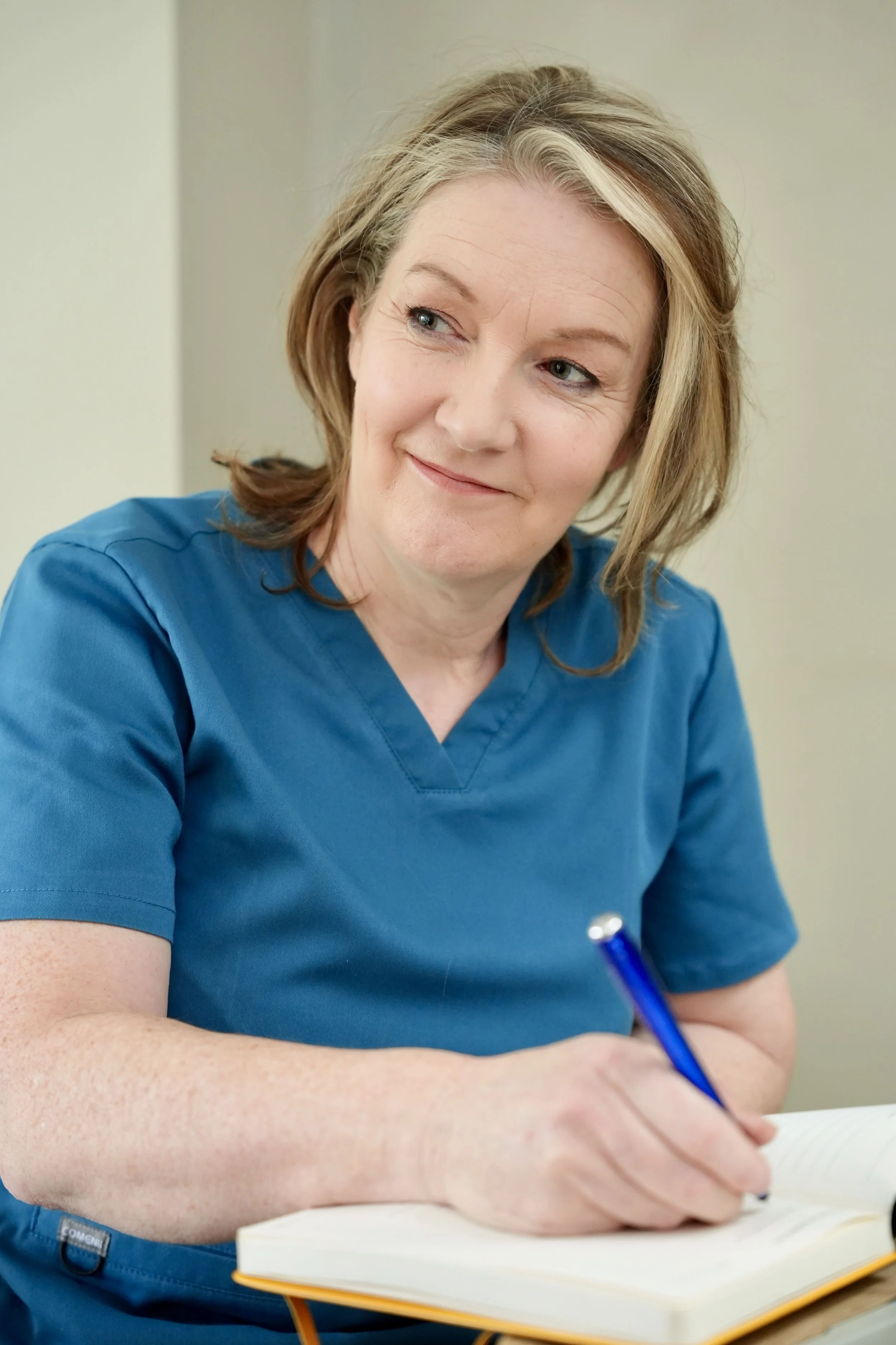 A woman with shoulder-length light brown hair wearing blue scrubs, sitting at a desk, writing in a notebook with a pen.