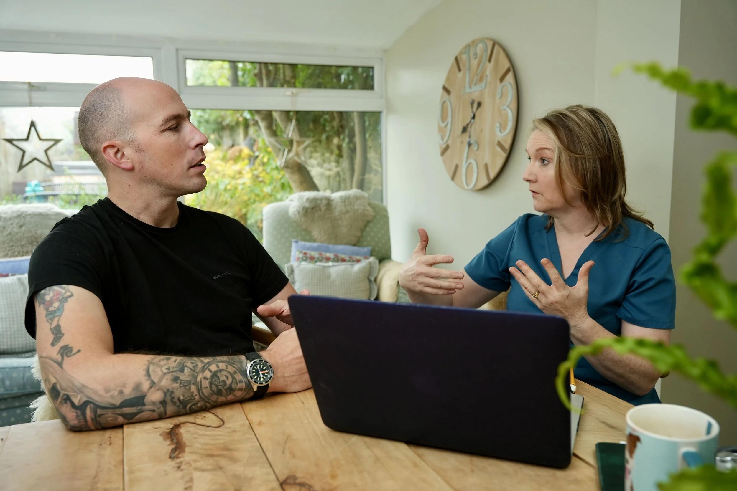 A man with tattoos on his arms and a woman wearing a blue shirt are sitting at a wooden table engaged in a conversation, with a laptop and coffee mugs on the table, in a room with large windows and a wall clock.