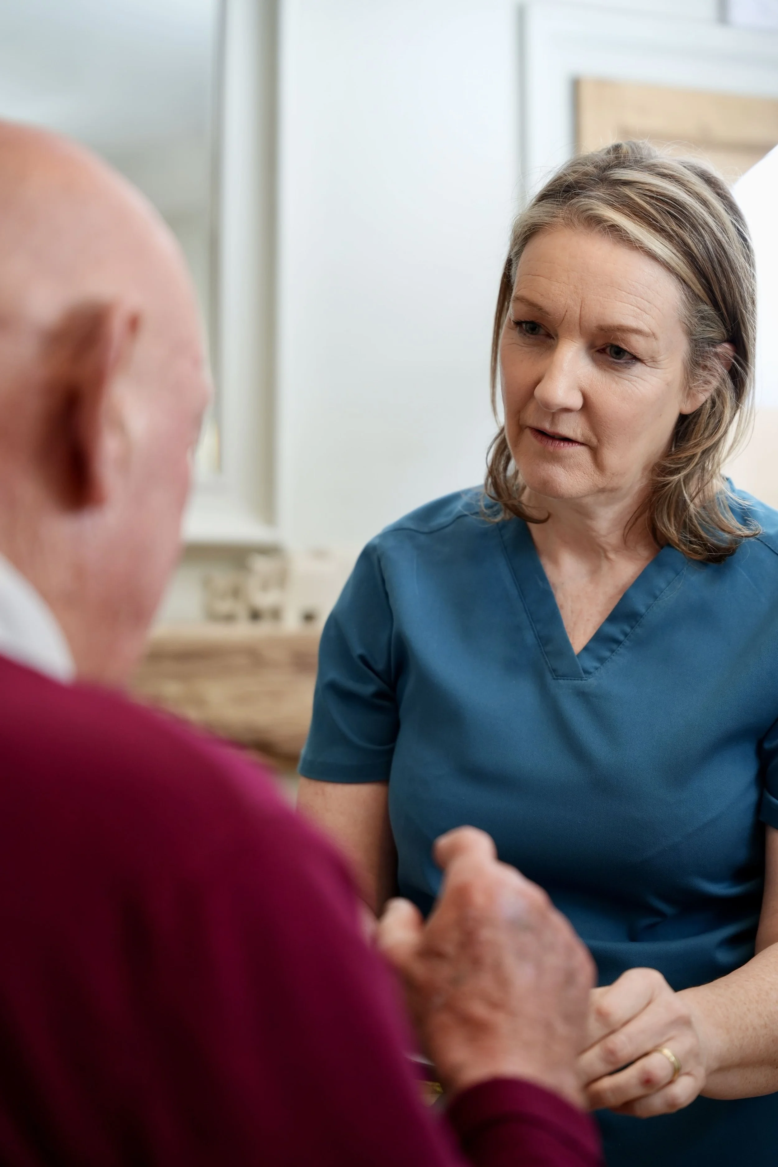 Cathy hearne, a female Osteopath in a blue uniform speaking with an elderly patient in a therapy room.