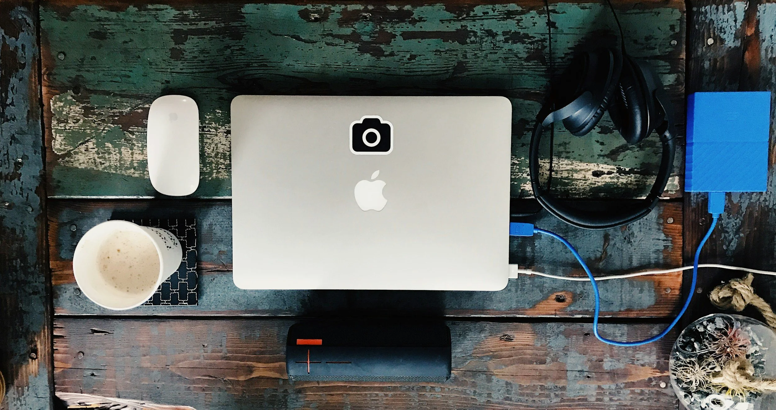 A wooden table with a MacBook, an Apple Magic Mouse, a cup of coffee, headphones, a portable speaker, a blue external hard drive, and some dried plants.