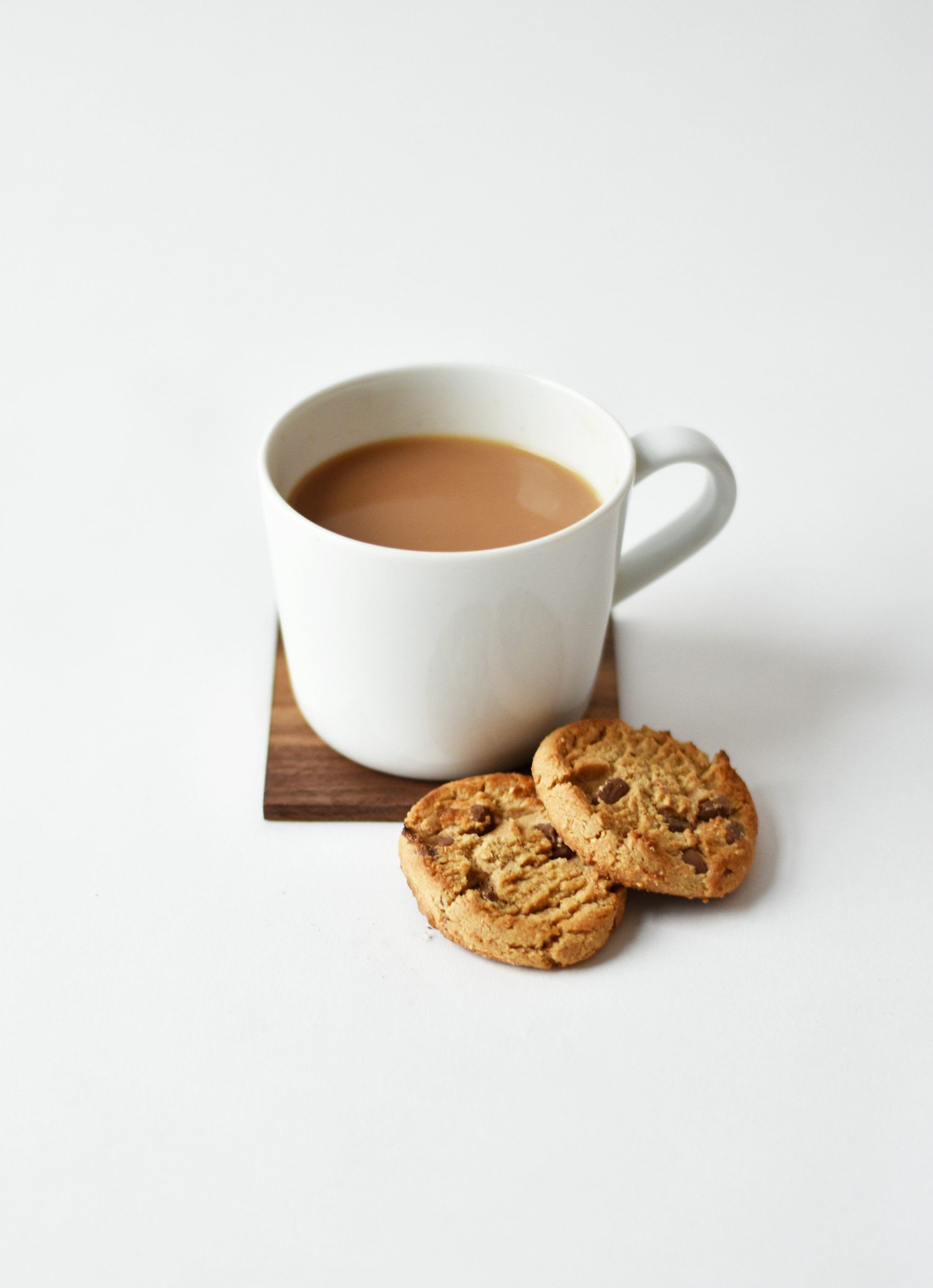 A white mug of coffee or tea on a wooden coaster with two chocolate chip cookies on a white surface.
