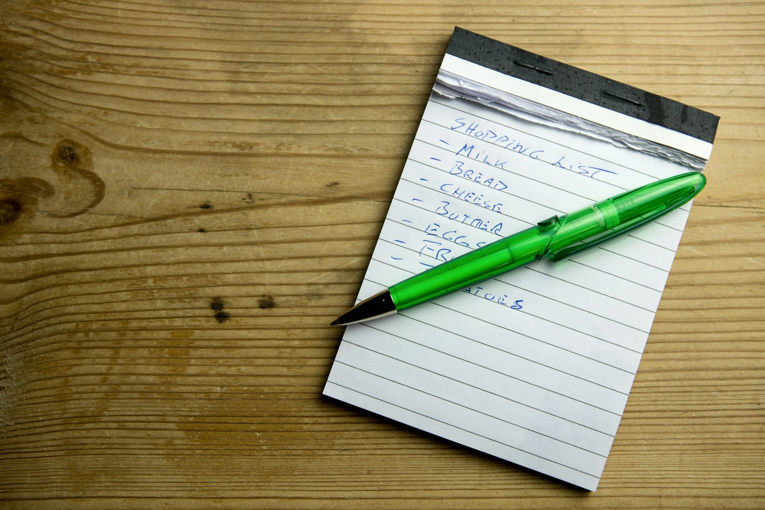 A notepad with a shopping list written in blue ink, placed on a wooden surface with a green pen resting on top.