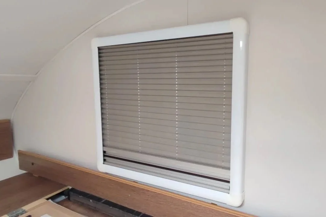 A window with closed beige horizontal blinds inside a white frame, on a wall above a wooden headboard.