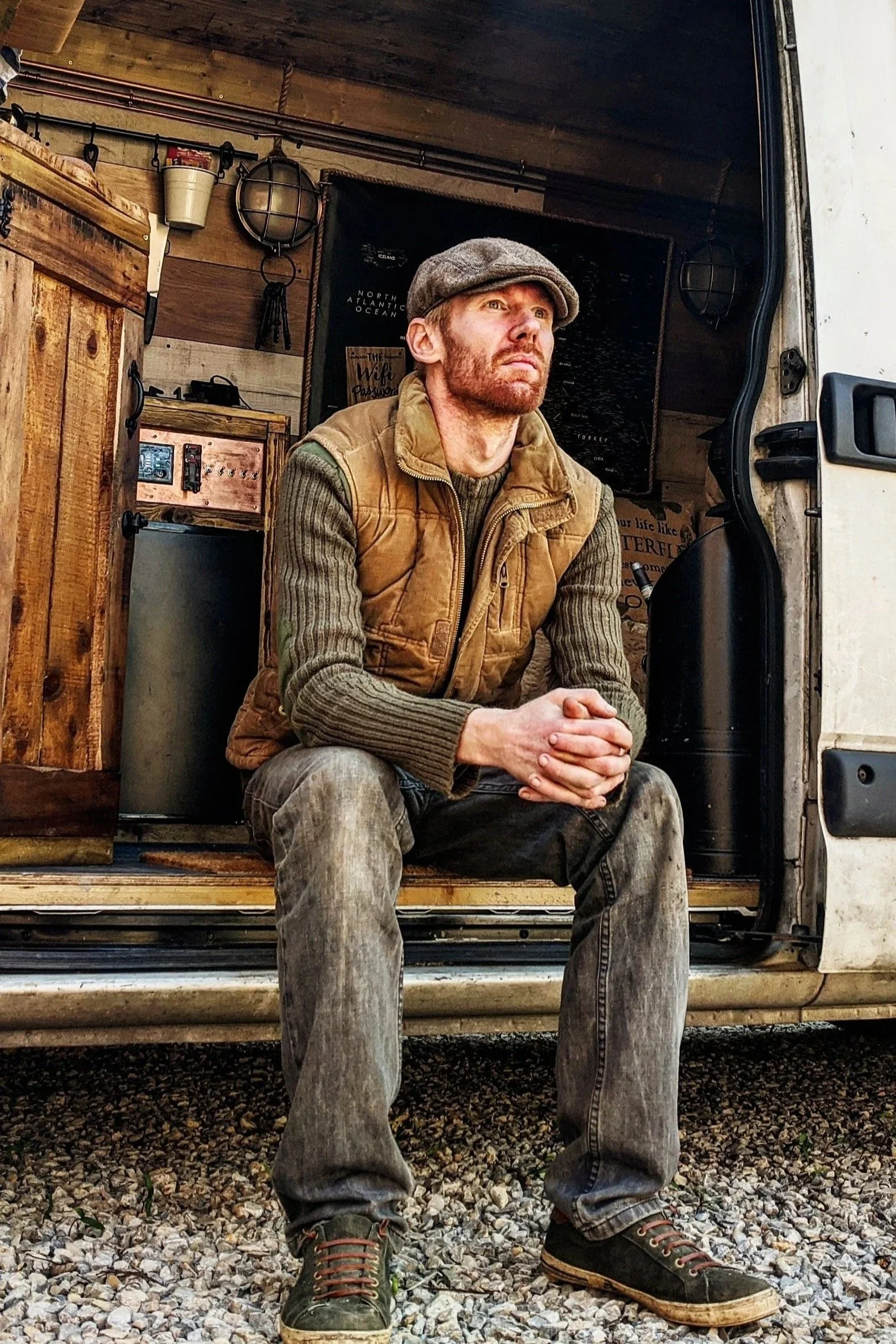 Man sitting on the edge of an open vehicle with wooden interior, wearing a cap, vest, and sweater, looking contemplative.