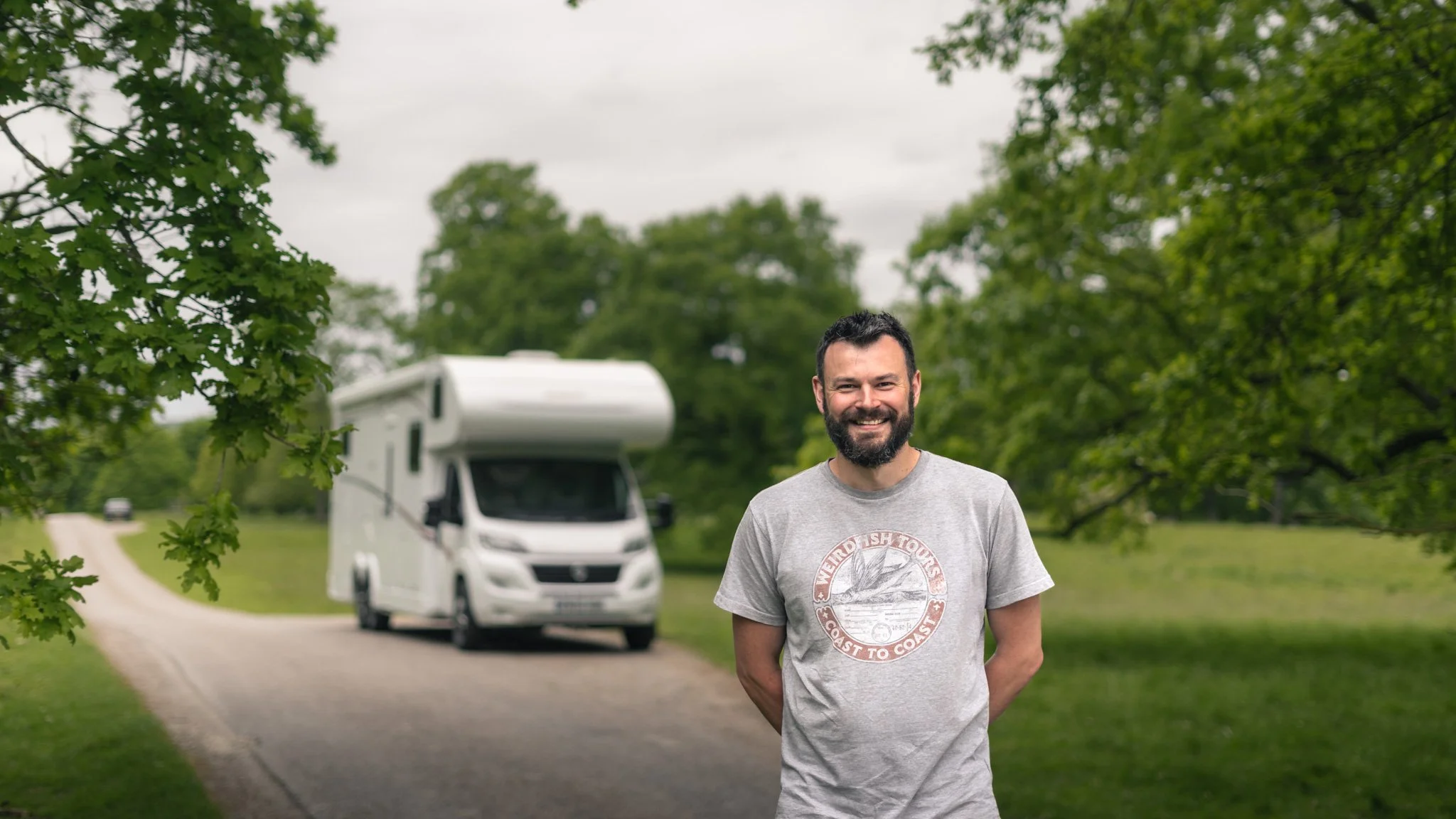 A bearded man wearing a gray T-shirt standing outdoors on a gravel path, with a camper van behind him and green trees on either side.