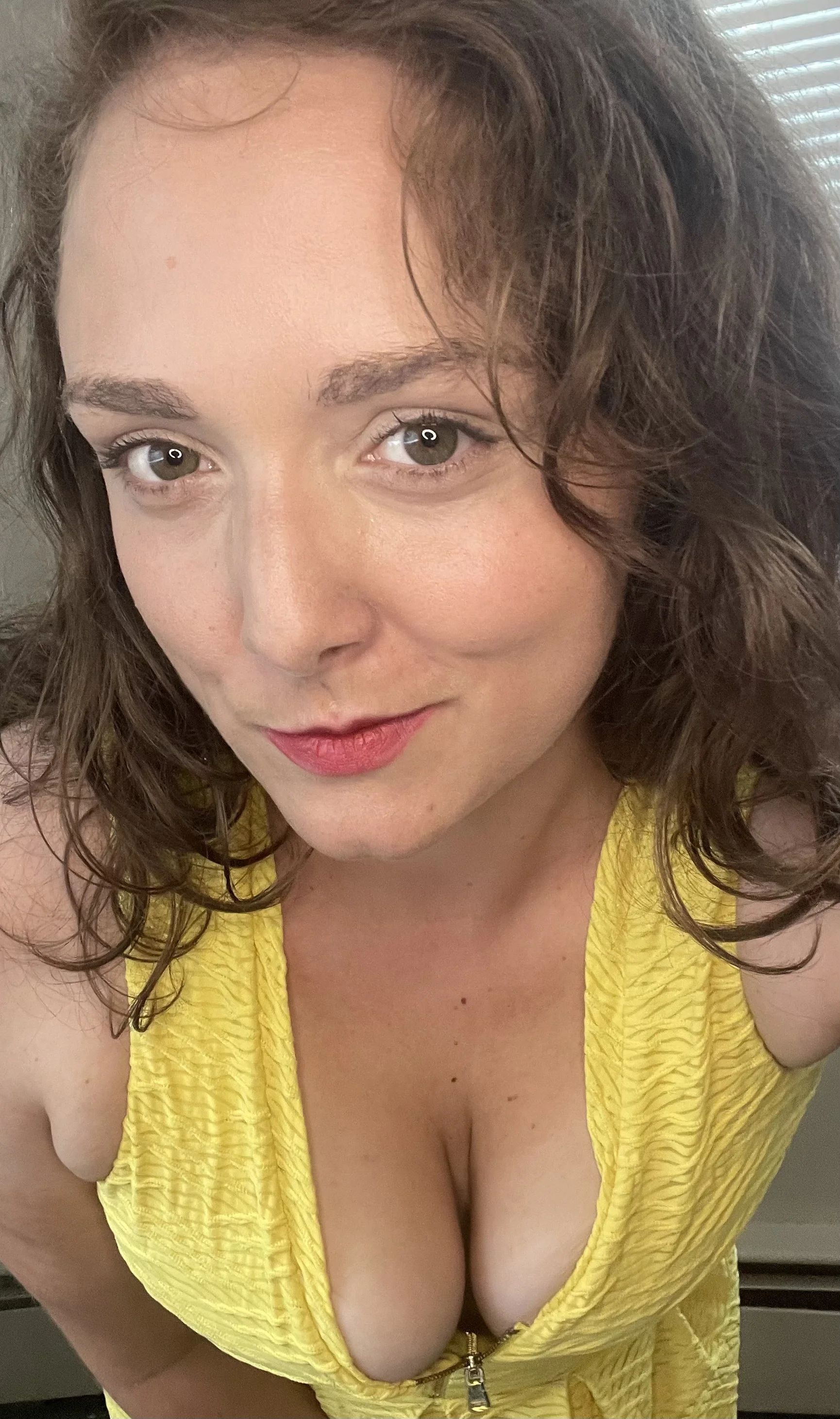 Close-up of a woman with wavy brown hair and fair skin wearing a yellow top with a zipper, smiling softly at the camera.