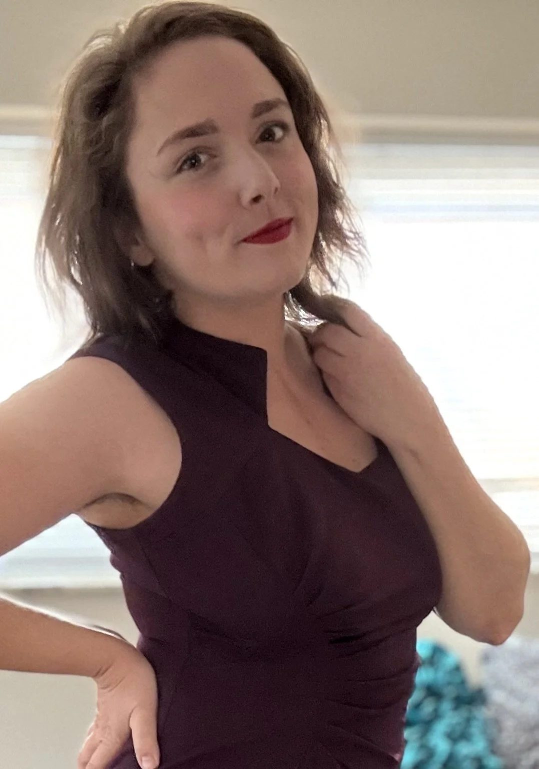 A woman with short, wavy brown hair and red lipstick, wearing a sleeveless dark purple dress, standing indoors in front of window blinds, with one hand on her waist and the other near her neck, smiling softly at the camera.