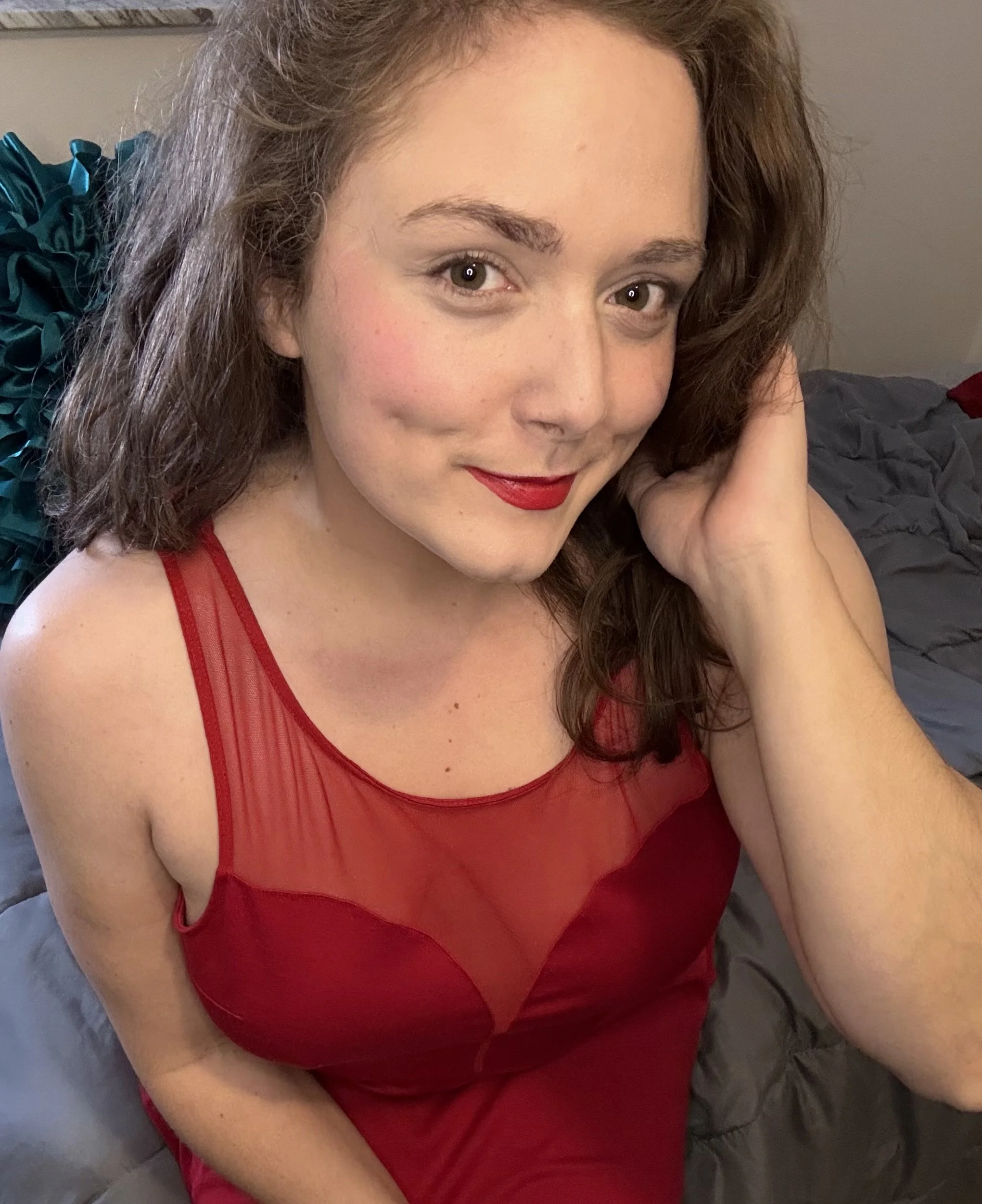 A woman with light skin and brown hair wearing a red dress with sheer detailing, posing indoors with a slight smile.
