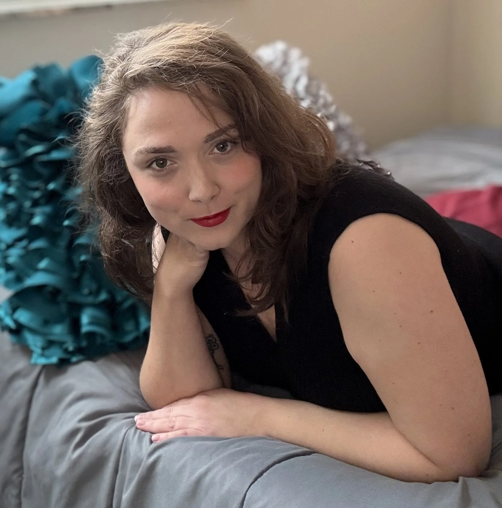 A woman with brown hair and red lipstick lying on a bed with a turquoise ruffled pillow and a gray comforter, resting her head on her hand and looking at the camera.