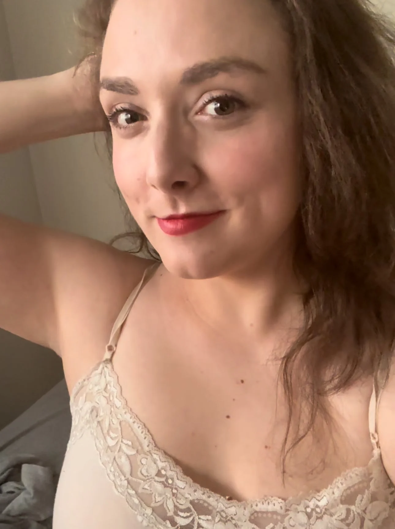 Close-up of a woman with brown curly hair, wearing a beige lace top, smiling with red lipstick, and resting her hand on her head.